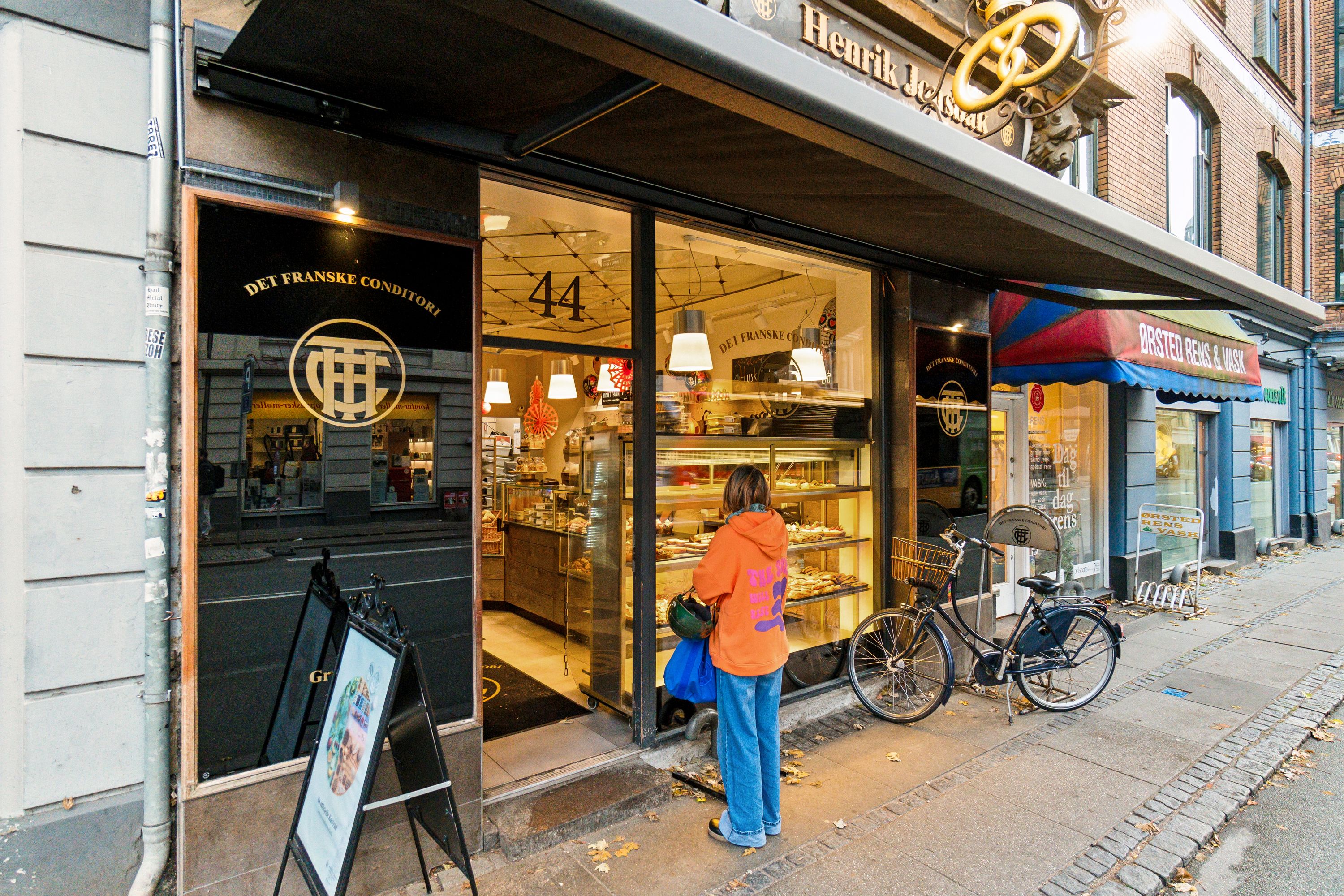 Kvinde i orange trøje og jeans står på gågaden og ser ind i udskærmet vindue på Det Franske Conditori på H.C. Ørsteds Vej 44