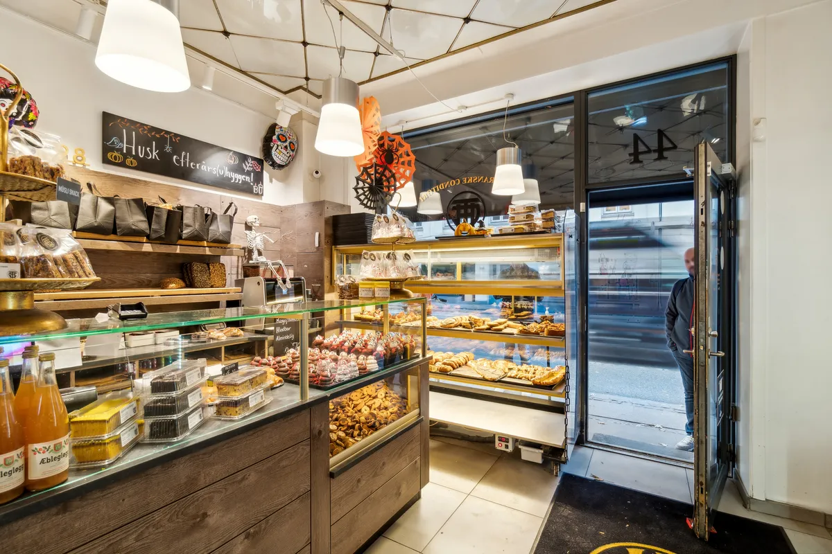 En salgsdame står ved indgangen til bageriet og vender sig mod kunden. Glasskurven indeholder forskellige kager og cookies. Der er flere træhylder med snacks og slik