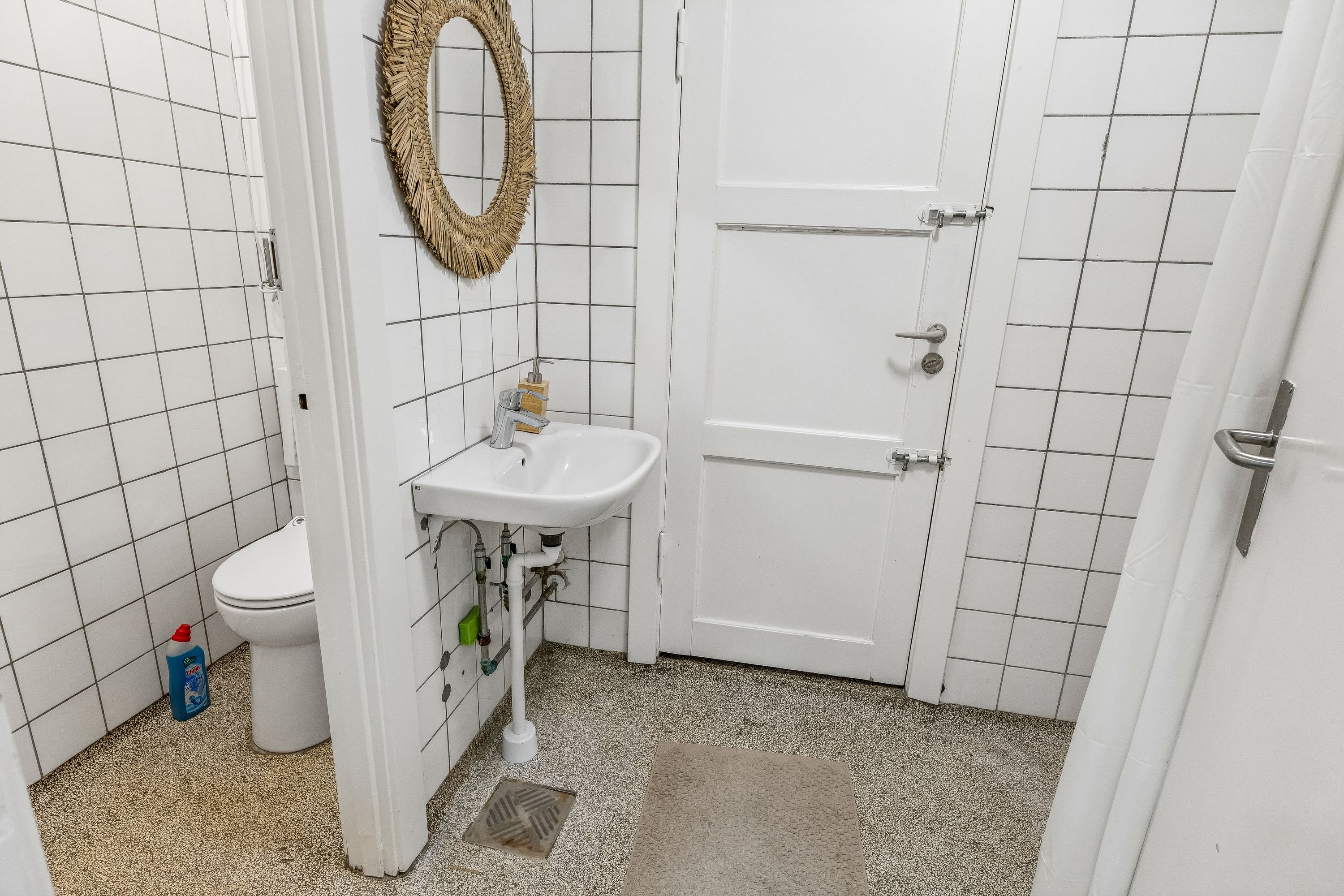 På et lille badeværelse med hvidt loft og hvide fliser er der en hvid toilet, hvid vask og en hængende spejl med ramme af strø