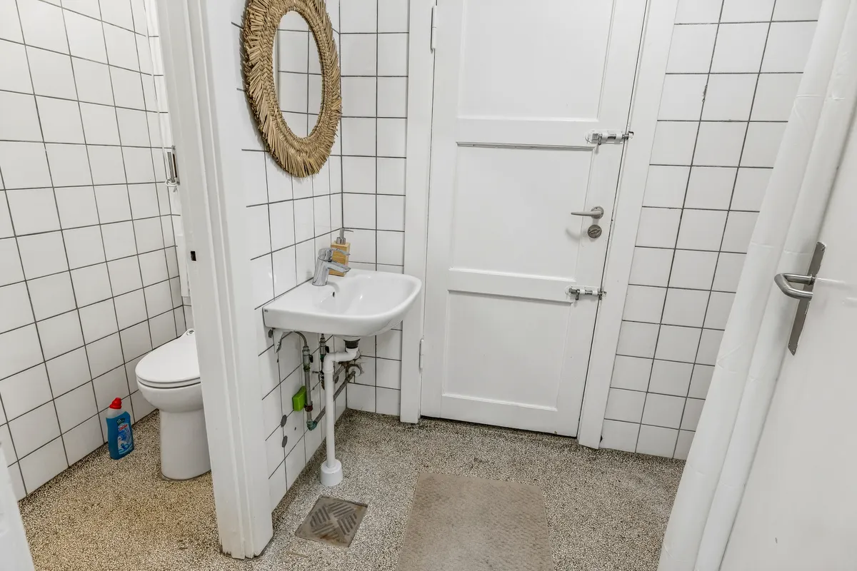 På et lille badeværelse med hvidt loft og hvide fliser er der en hvid toilet, hvid vask og en hængende spejl med ramme af strø