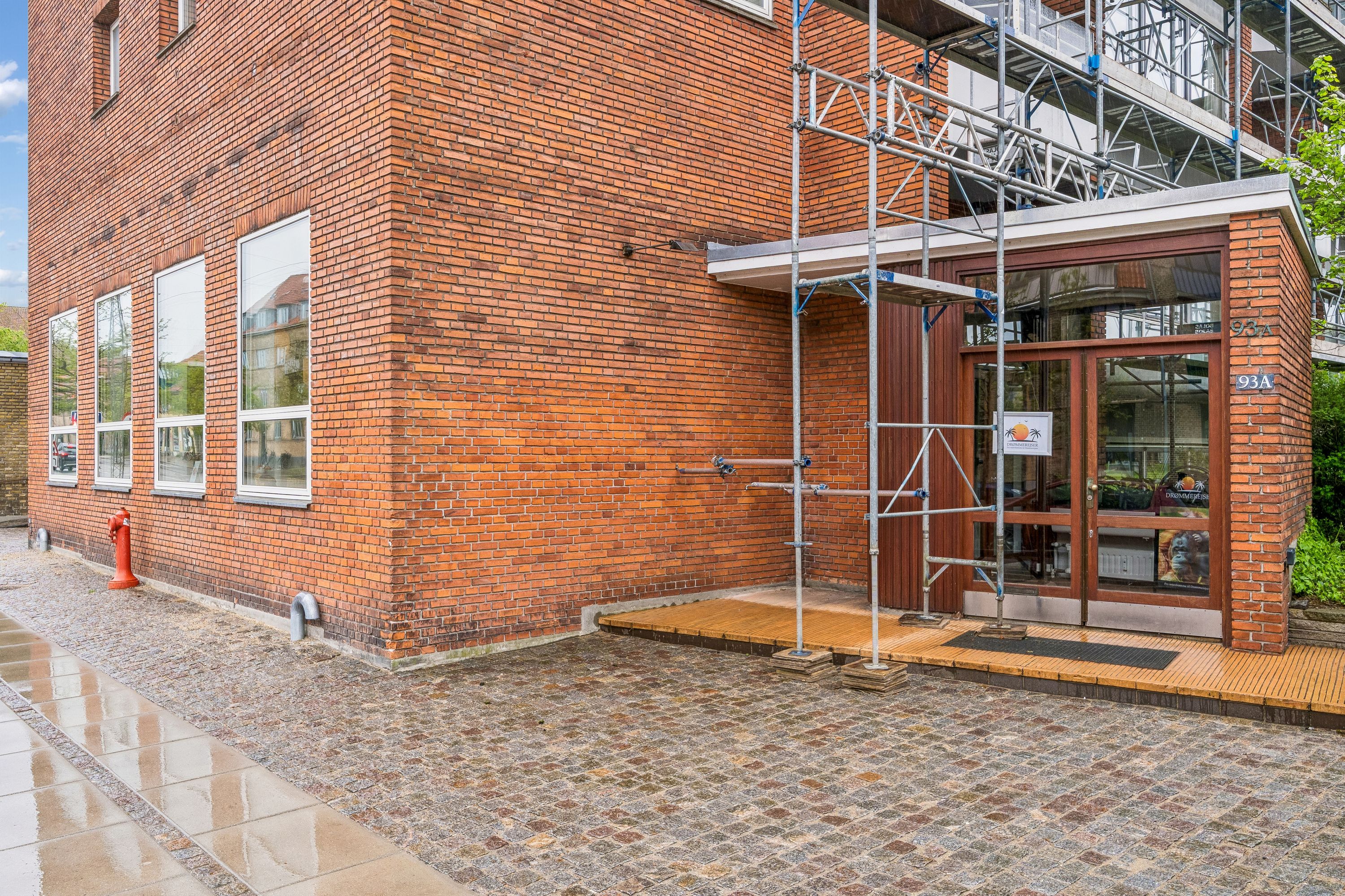 Kontor til leje på Godthåbsvej 93A, Frederiksberg. Stenbrolægget fortov med rødt murstenshus og glasvinduer. Skafte og rødt brandvandshus. Opførte stelninger og vinduer.