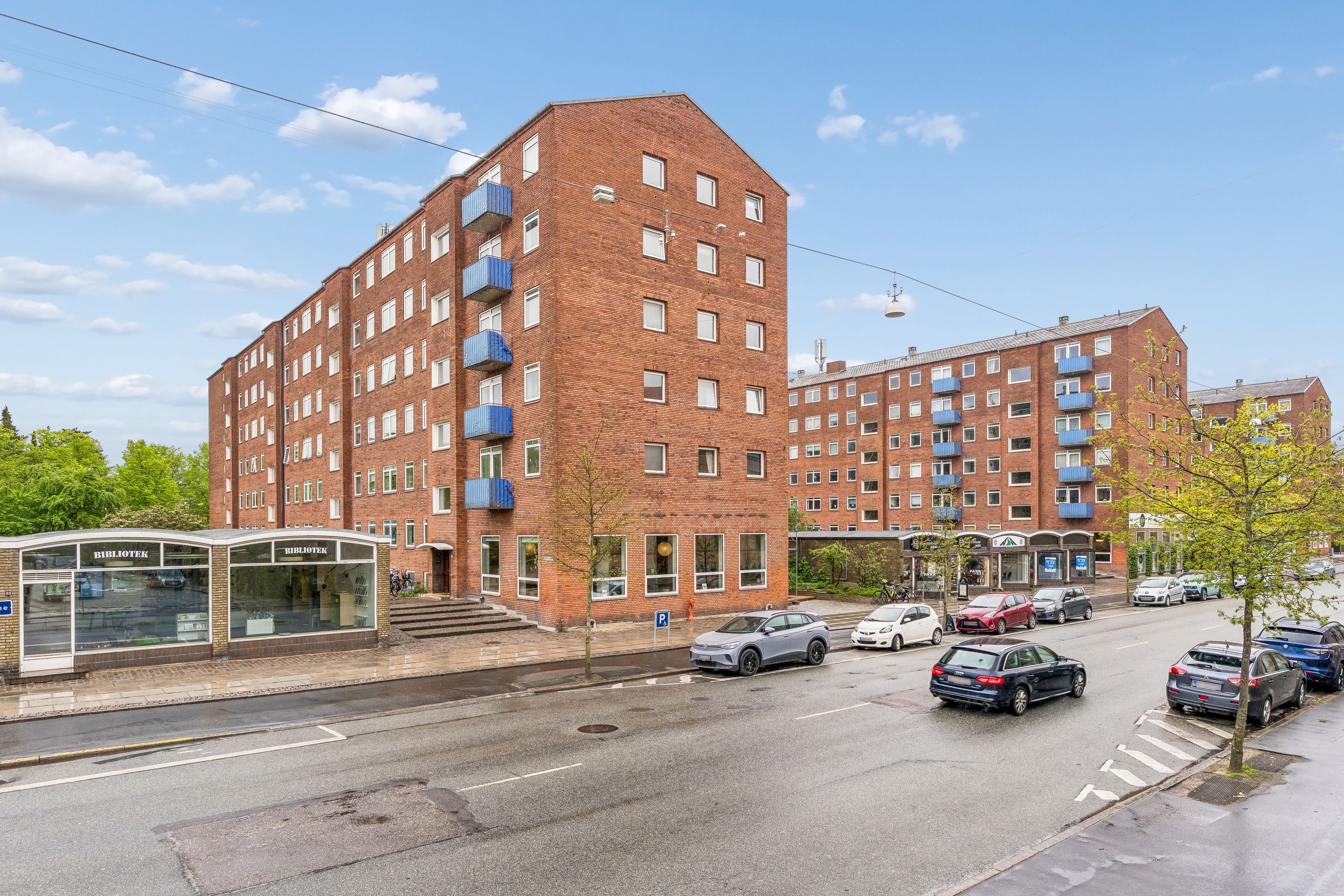 Stående kontorer med facade i rød mursten. St. 1 på Godthåbsvej 93A. Parkerede biler på højre side af vejen. Bibliotek og forretninger med trapper og balkoner