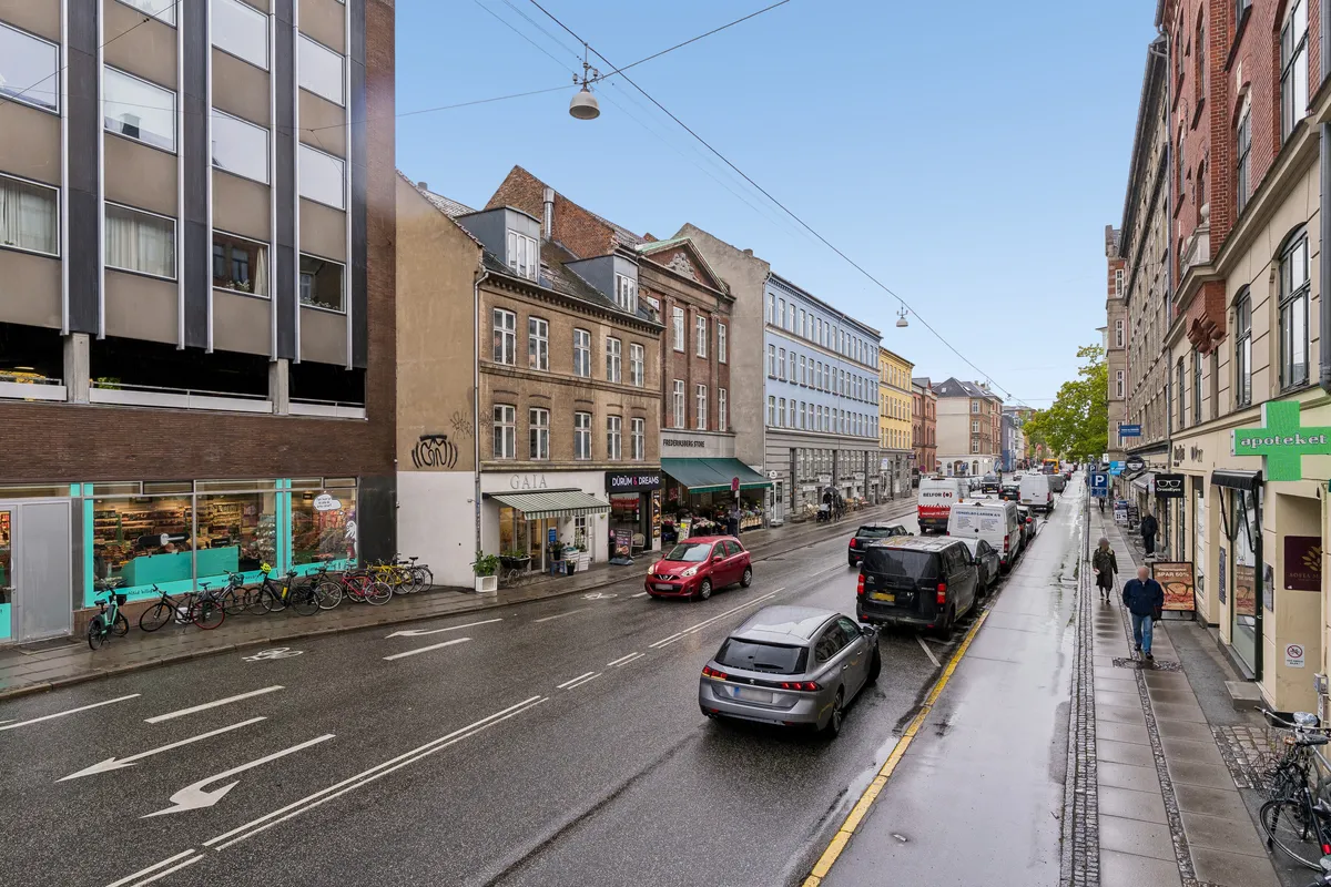 Butiksgade med parkerede biler, cykler og gående folk i Frederiksberg. Kæde af bygninger med vinduer, skilte og grønne vinduer. Vej med skilte og busstop.
