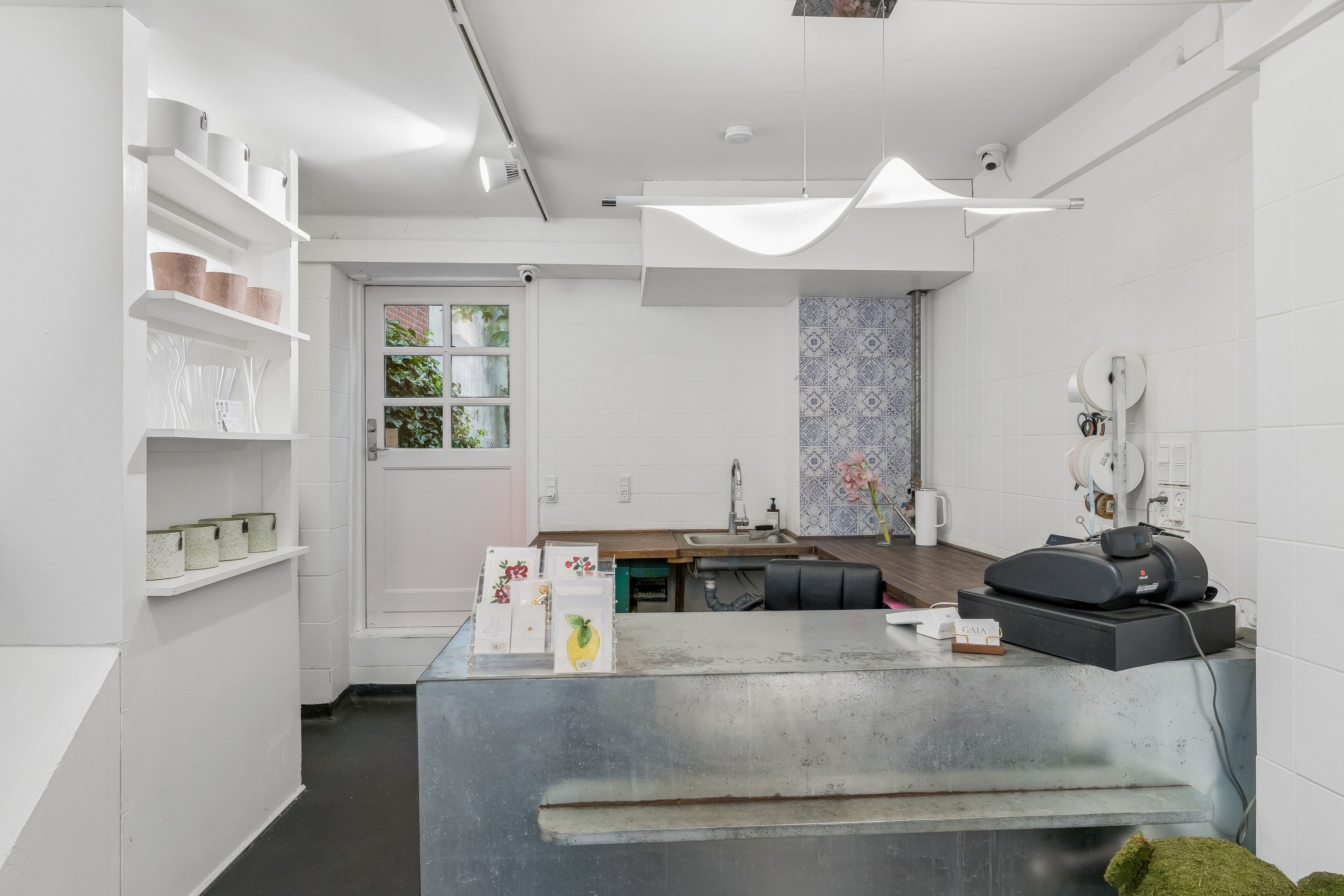 Butiksrum med hvid væg og gulv. Tæller med blomster og papirkurve. Komplet med metalbord og elektronisk salgssystem. Der er et hvidt glasvindue med udsigt til have.