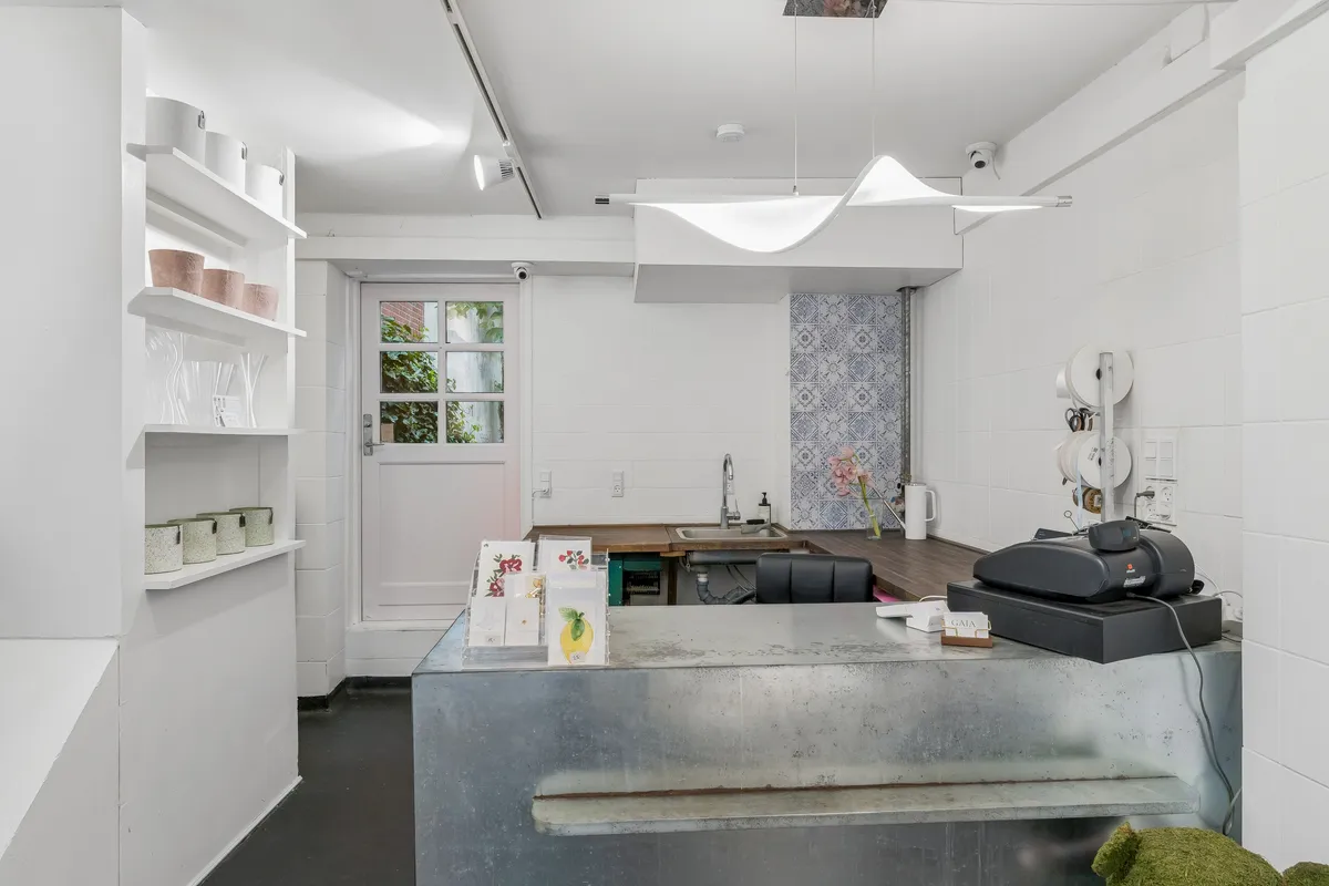 Butiksrum med hvid væg og gulv. Tæller med blomster og papirkurve. Komplet med metalbord og elektronisk salgssystem. Der er et hvidt glasvindue med udsigt til have.