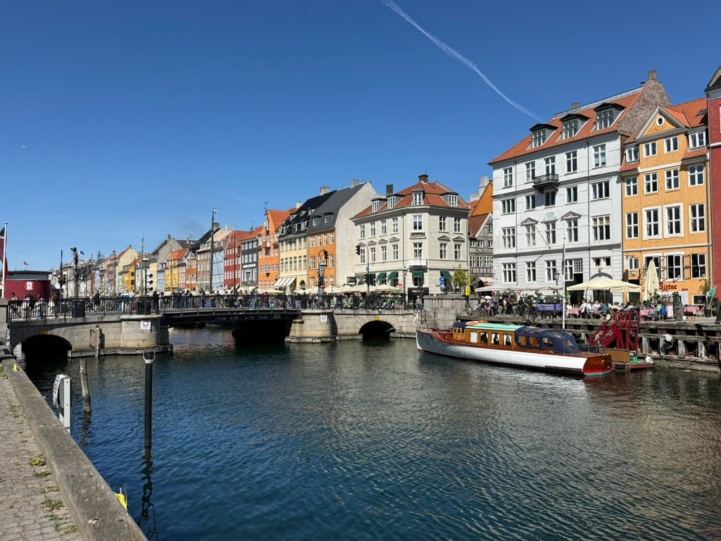 Kontor i Nyhavn med udsigt til vandet og flere bygninger og en båd. Her er flere turister og folk går på gaden og står foran bygningerne