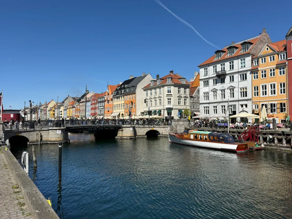 Kontor i Nyhavn med udsigt til vandet og flere bygninger og en båd. Her er flere turister og folk går på gaden og står foran bygningerne