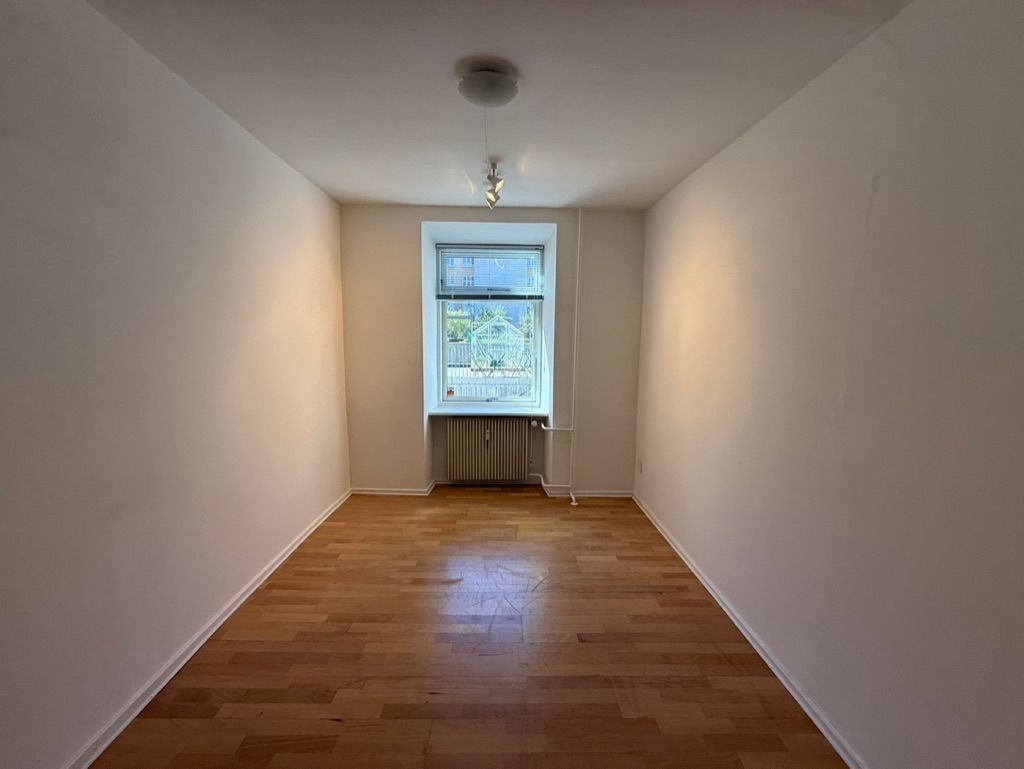 Et fladt rum med lyse, hvide vægge og en trægulv. Der er et vindue med udsigt til en bygning og et træ. Der er et radiator i hjørnet