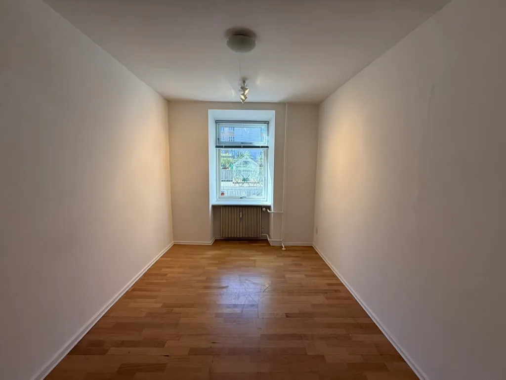 Et fladt rum med lyse, hvide vægge og en trægulv. Der er et vindue med udsigt til en bygning og et træ. Der er et radiator i hjørnet