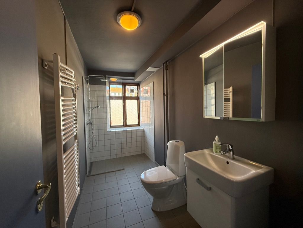 Rent badeværelse med en hvid toilet, hvidt badekar og hvidt spejl med indbygget lys. Der er en hængelampe og en sæbeflaske på vasken