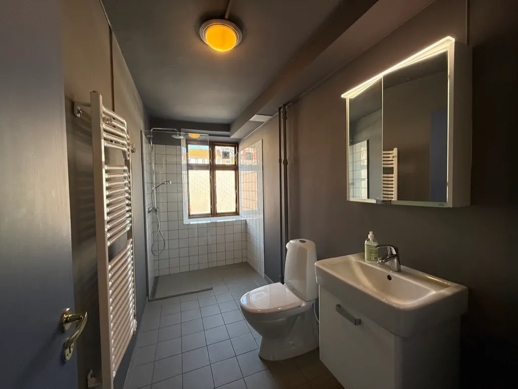 Rent badeværelse med en hvid toilet, hvidt badekar og hvidt spejl med indbygget lys. Der er en hængelampe og en sæbeflaske på vasken