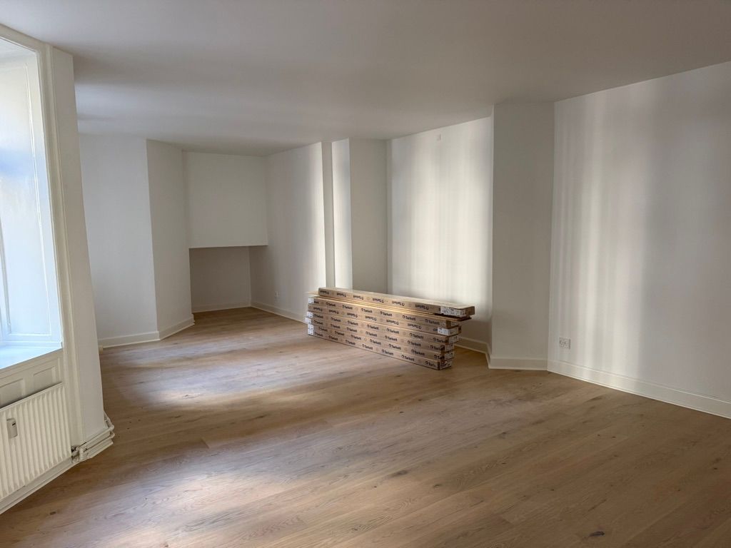 En tom kontorlokal med hvid malet væg, trægulv og gulvbelægning i pakket stel. Et hvidt radiator står mod væggen