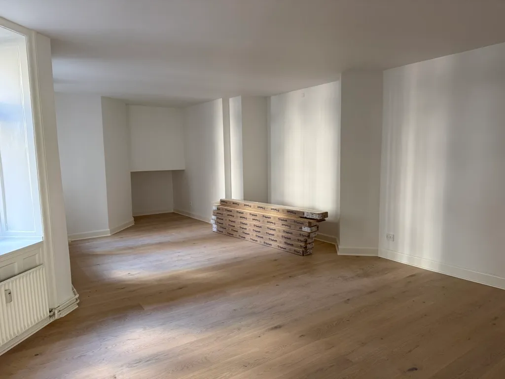 En tom kontorlokal med hvid malet væg, trægulv og gulvbelægning i pakket stel. Et hvidt radiator står mod væggen