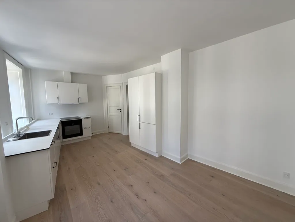 En møbleret køkken med hvide vægge og loft samt højloftede rum, hvor der er en spisestue, ovn og en indbygget køleskab.