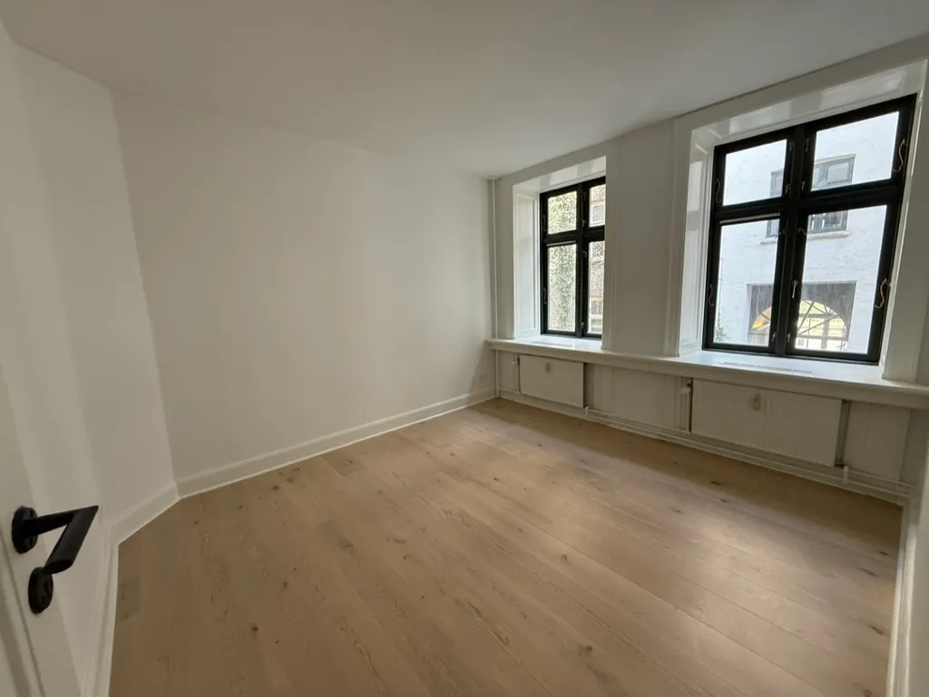 Tomt kontor med hvide vægge og gulvtæppe, to store vinduer med lysegardiner, moderne radiatorer og et sort håndtag på døren