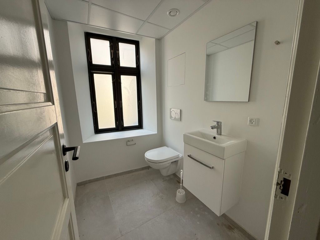 Rent og hvidt toiletrum med toilet, hvidt vask og spejl samt et vindue med udsigt til gaden