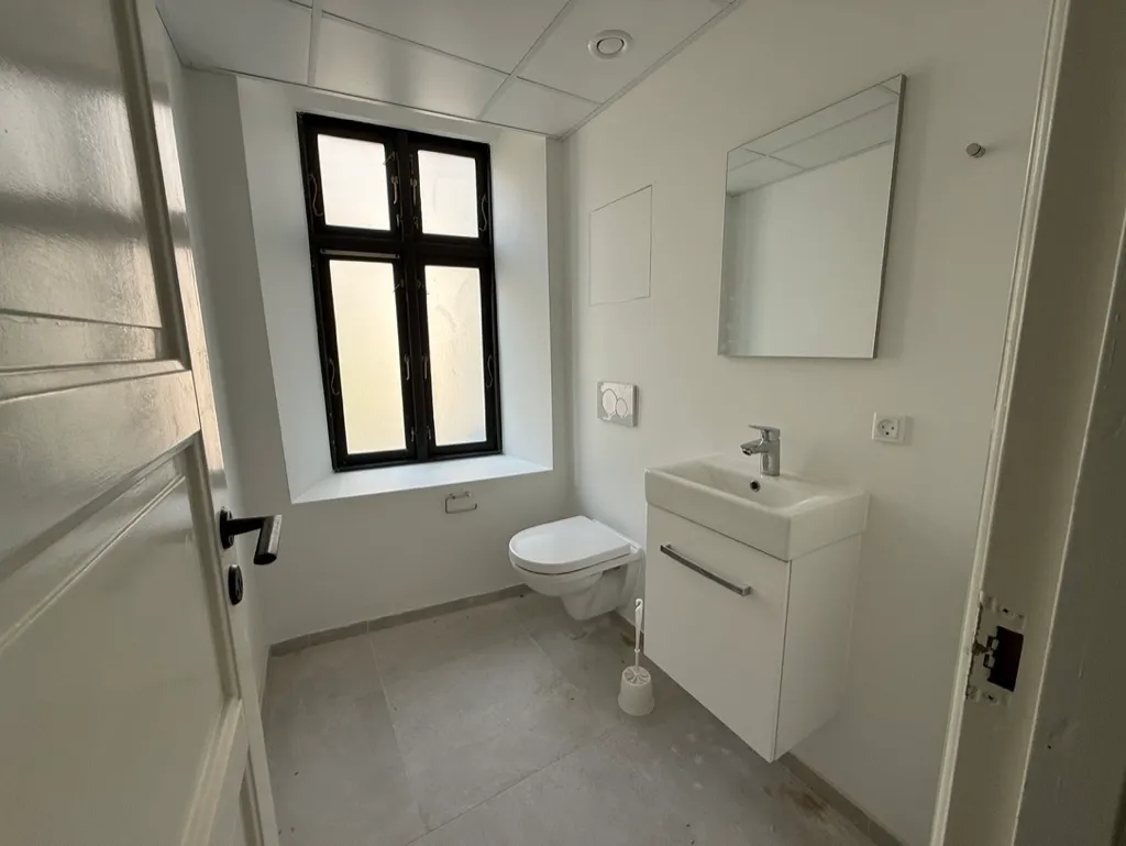 Rent og hvidt toiletrum med toilet, hvidt vask og spejl samt et vindue med udsigt til gaden