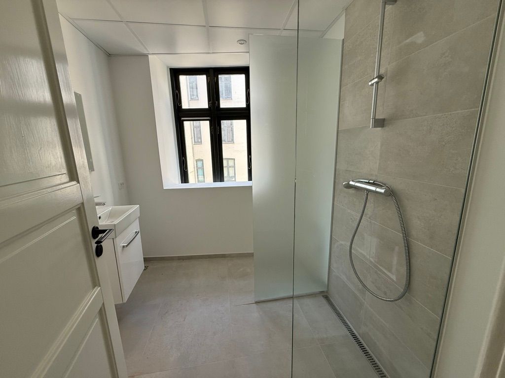 En moderne badeværelse med glasdør, hvidt badekar og lysegule vægge og gulv samt en stor bueformet vinduesglas.