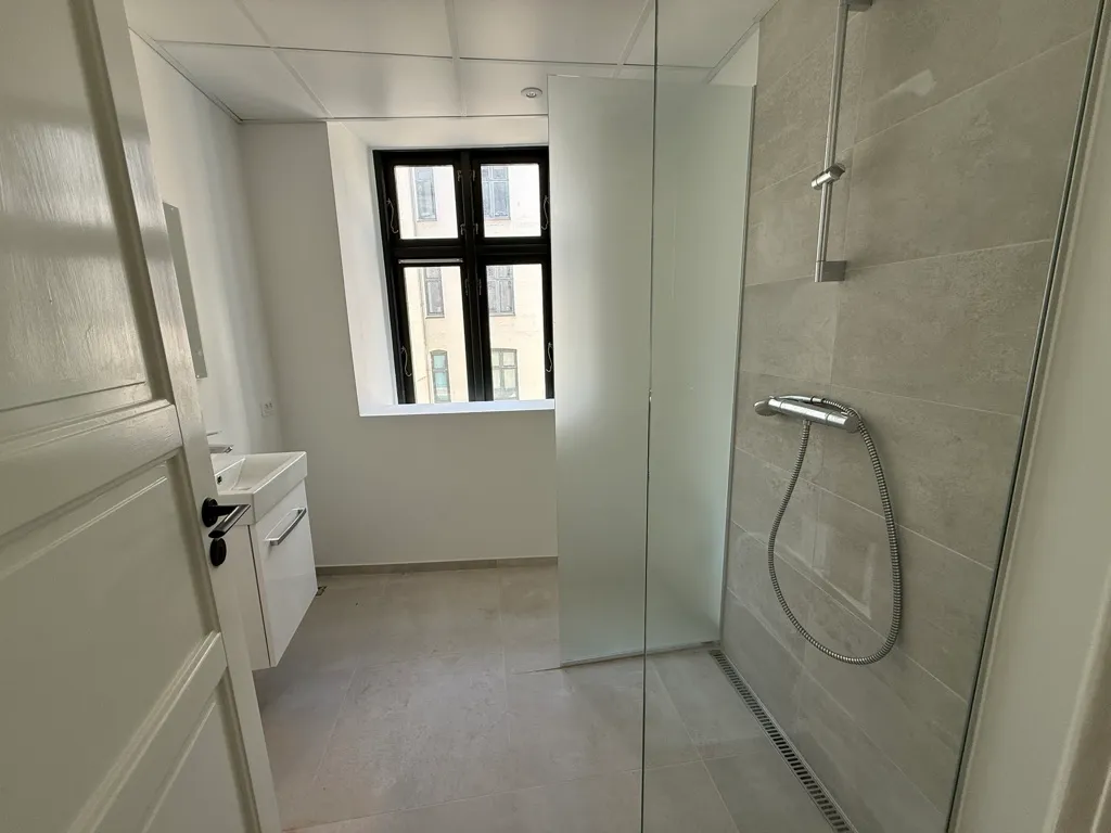 En moderne badeværelse med glasdør, hvidt badekar og lysegule vægge og gulv samt en stor bueformet vinduesglas.