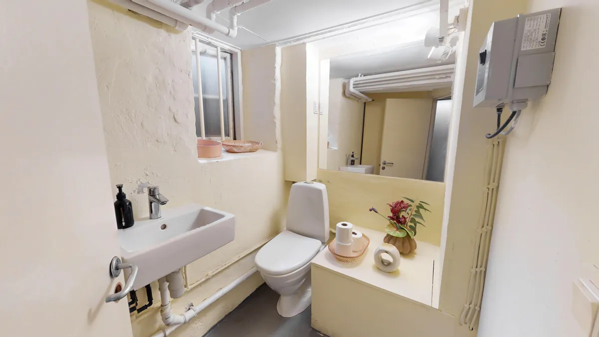 Rent og hyggeligt badeværelse med toilet, håndvask og spejl samt to toiletpapirruller og en vase med blomster.