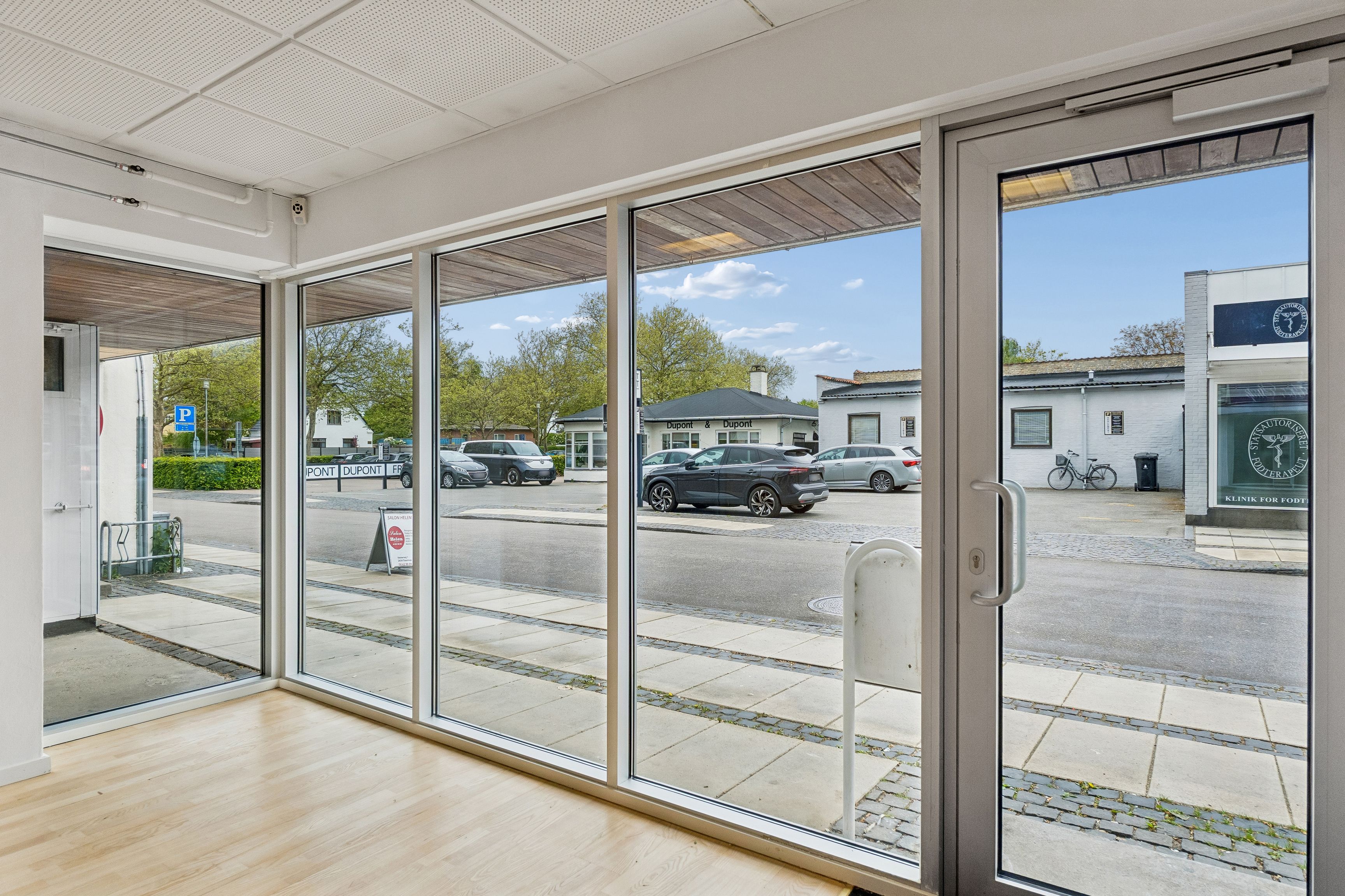 Lyst og stort butikslokale med store facadevinduer og synlig placering. Bag facaden er der moderne gulvbelægning og lyse lofter. Parkerede biler og grønne træer er synlige på gaden.