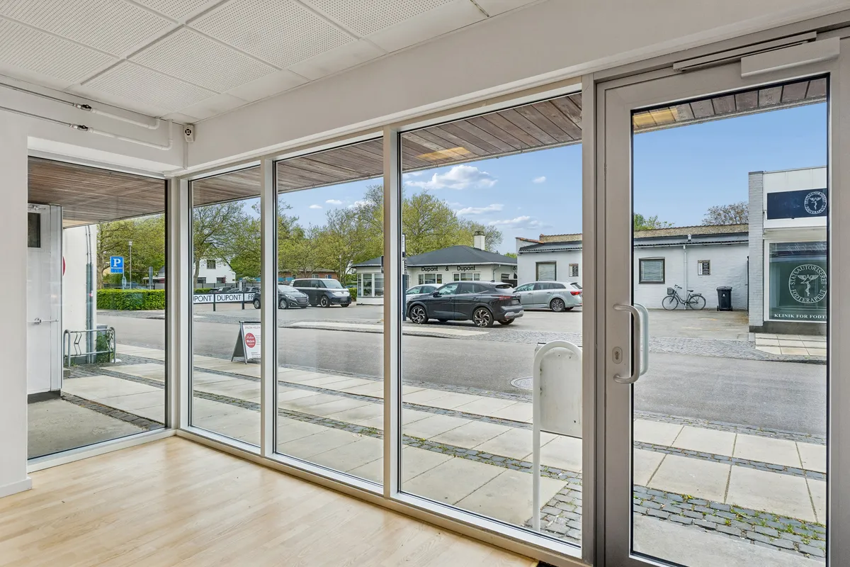 Lyst og stort butikslokale med store facadevinduer og synlig placering. Bag facaden er der moderne gulvbelægning og lyse lofter. Parkerede biler og grønne træer er synlige på gaden.