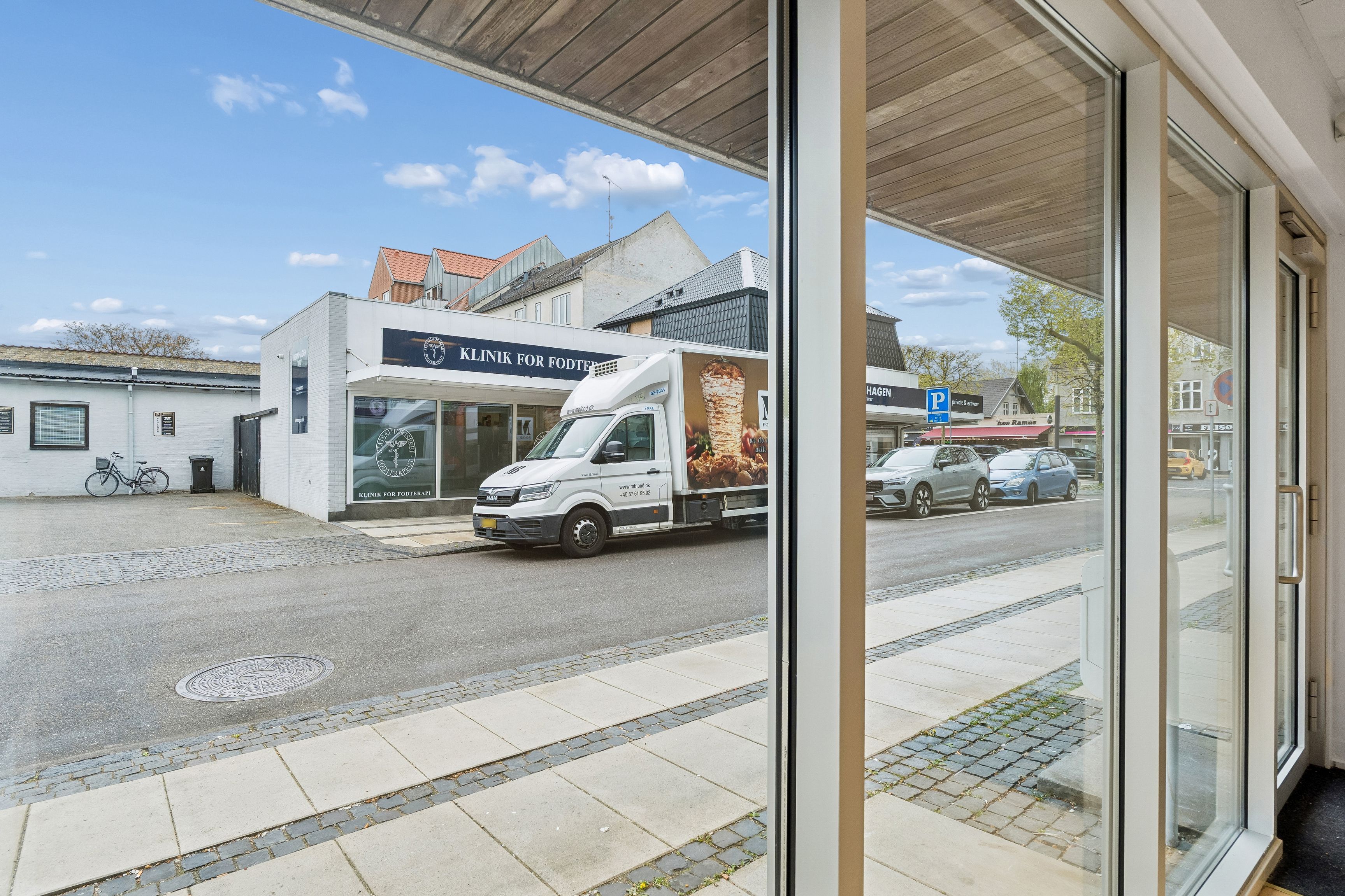 Lyst og charmerende erhvervslejemål med store facadevinduer. En hvid varevogn parkeret udenfor med reklame. To biler kører langs gaden. Solklar himmel og grønne træer.
