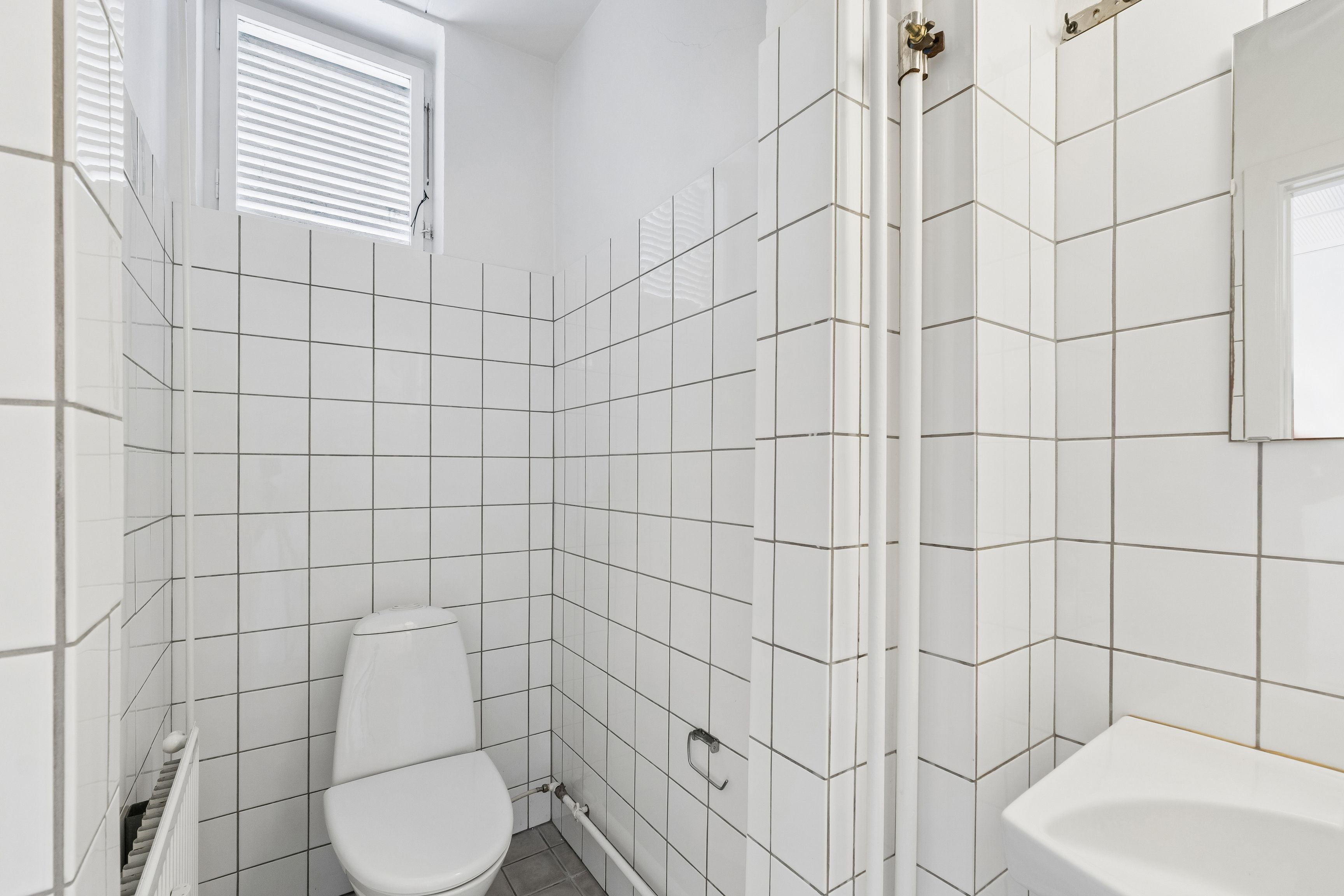 Lyst og moderne badeværelse med hvide kakel og toilet. Væggene og gulvet er klædt med hvide kakler, og der er et hvidt badekar med hvidt håndklæde.