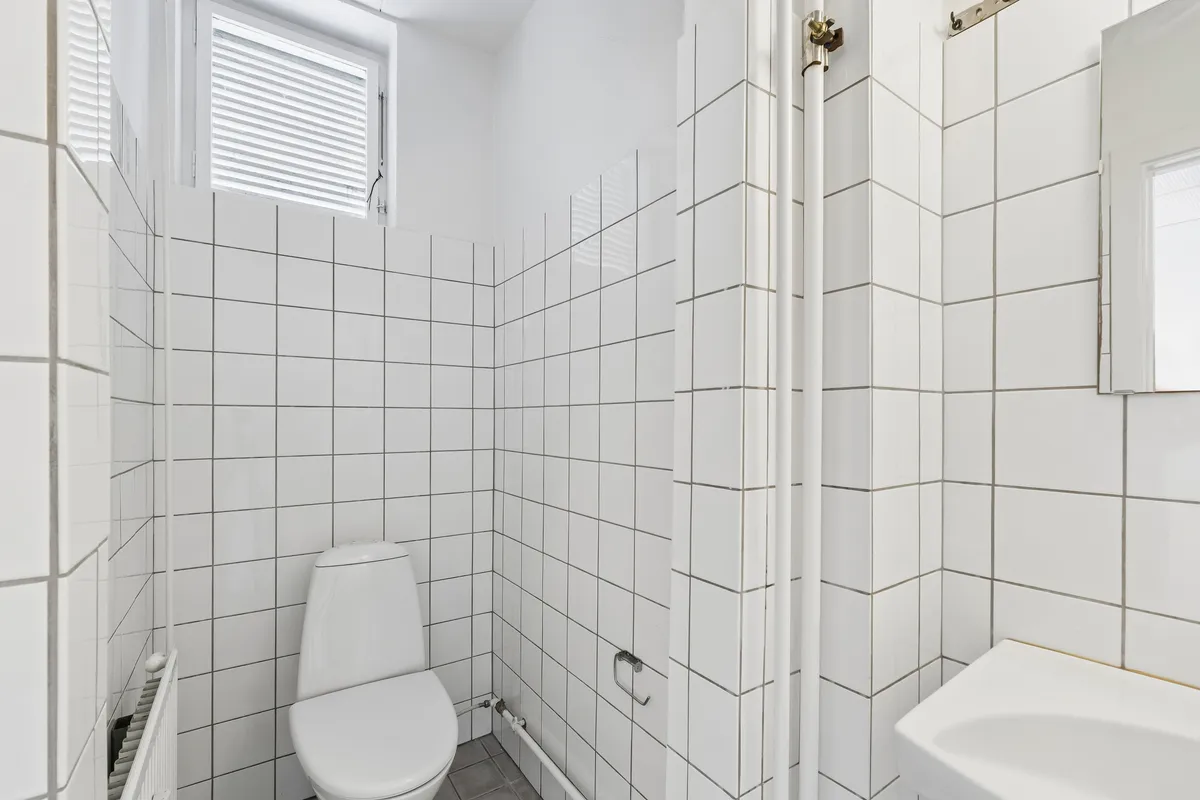 Lyst og moderne badeværelse med hvide kakel og toilet. Væggene og gulvet er klædt med hvide kakler, og der er et hvidt badekar med hvidt håndklæde.