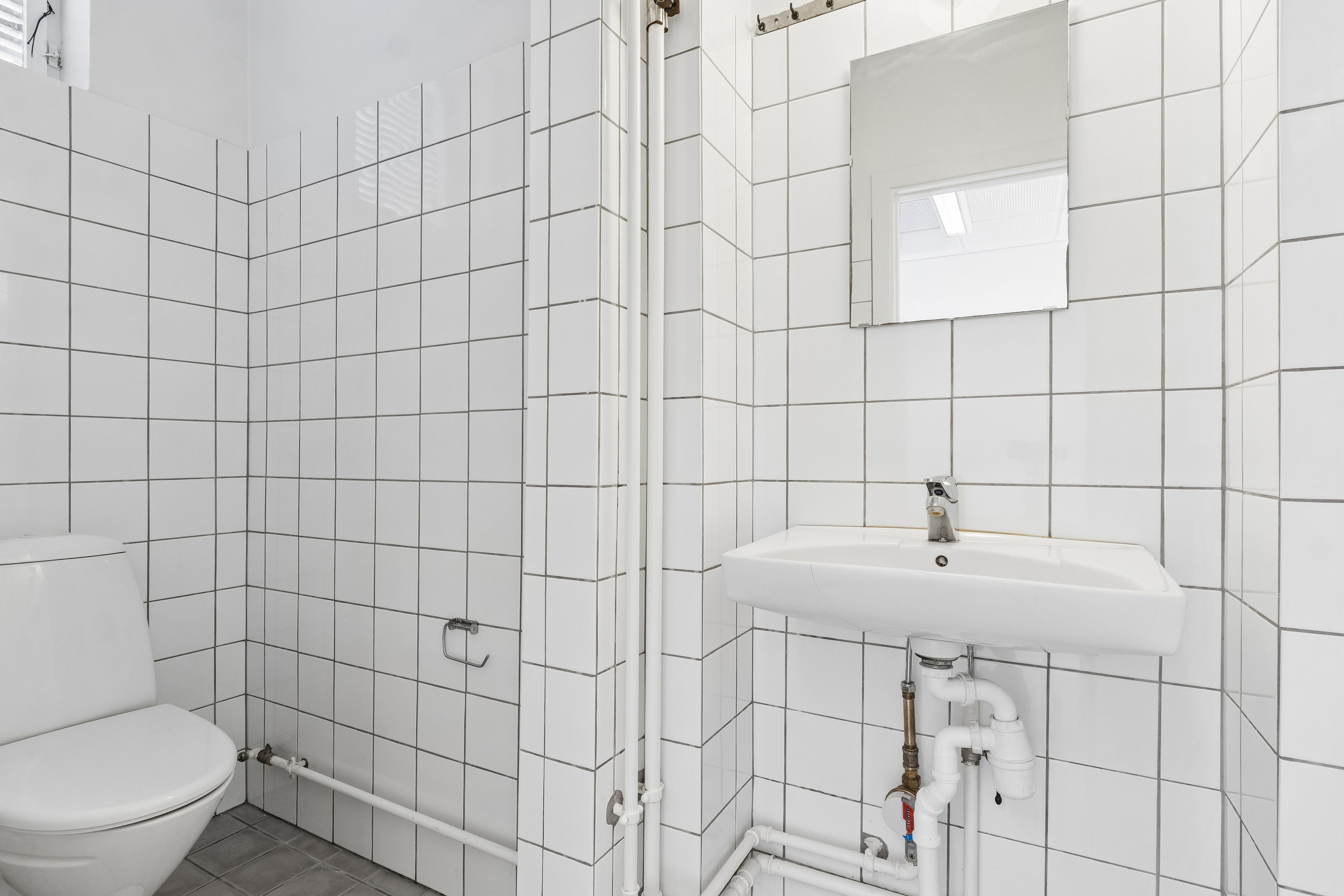 Lyst og rent toiletrum med hvide keramiske fliser og moderne design. Toiletstol og hvidt vasketøjsskab med håndklædeholder og spejl. Moderne renlighed og design i smukt og funktionelt toiletrum.