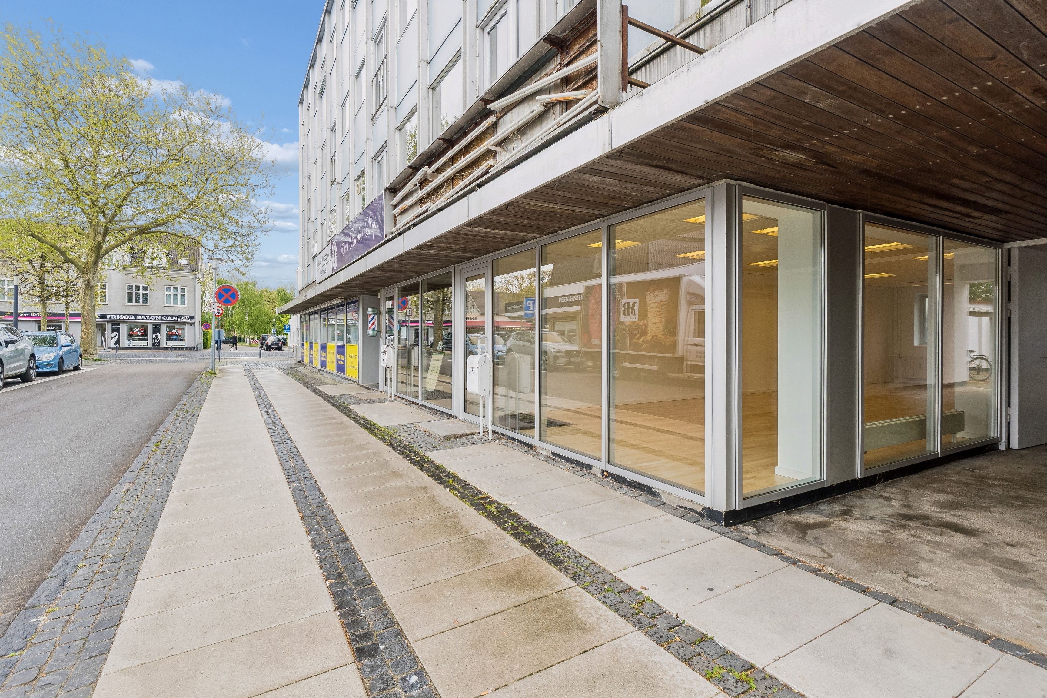 Lyst butikslokale med store glasvinduer og moderne facade. Beliggenhed med parkeringsplads og enkel adgang til gågaden. Solrig og attraktiv placering med let adgang til offentlig transport.
