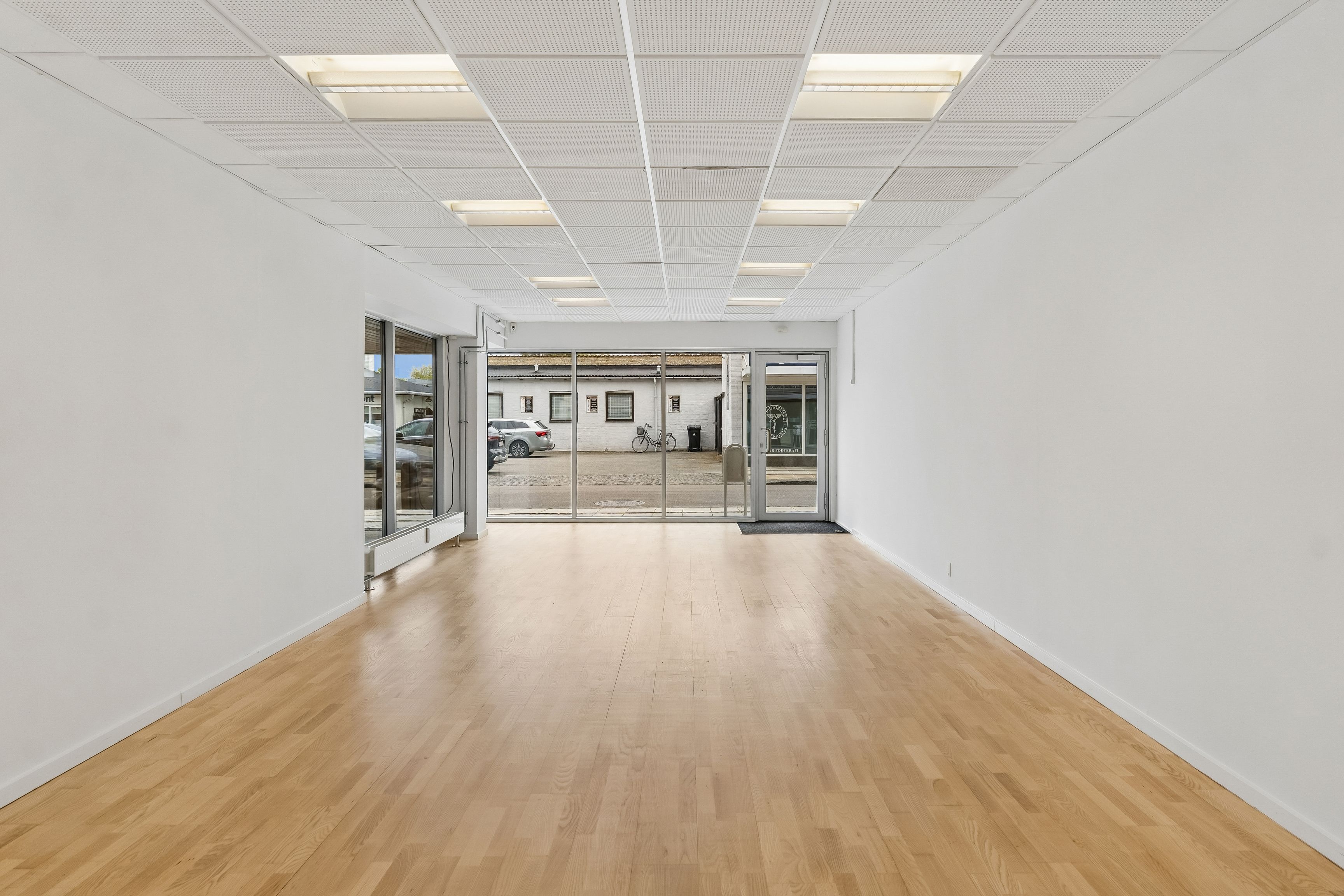 Lyst og åbent butikslokale med lyse loftslampe og store glasdøre. Lys og moderne design med massivt trægulv og lyse facadevinduer til stor synlighed.