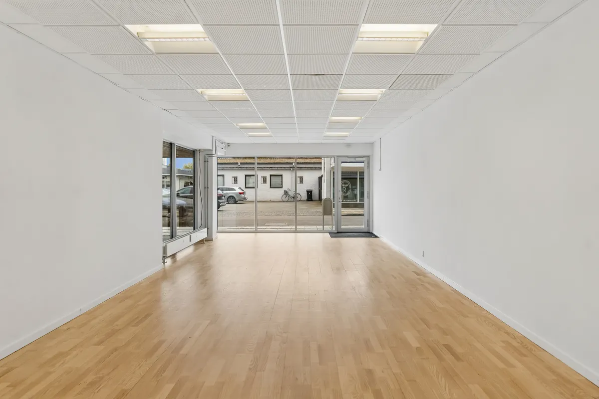 Lyst og åbent butikslokale med lyse loftslampe og store glasdøre. Lys og moderne design med massivt trægulv og lyse facadevinduer til stor synlighed.