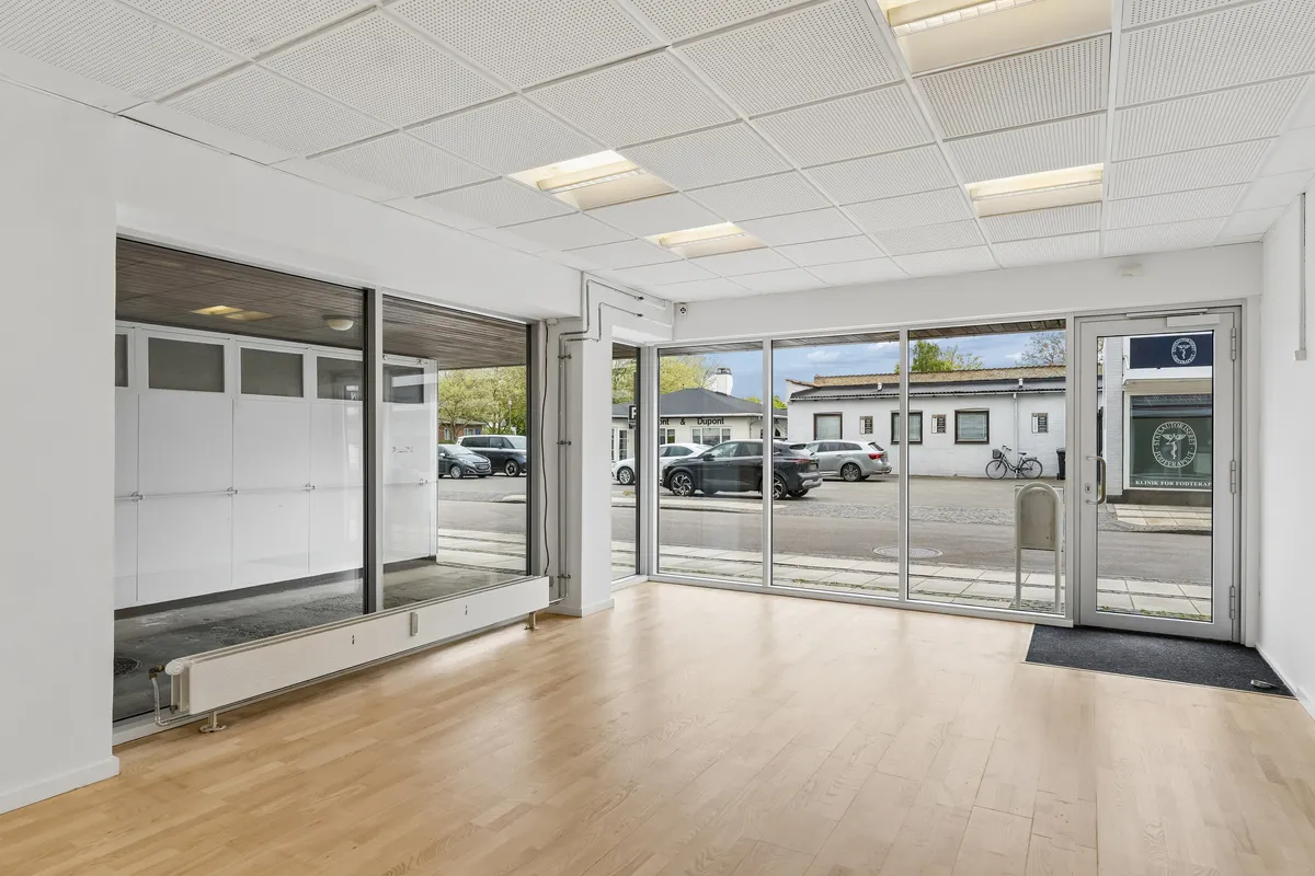 Store glasdøre og vinduer i hvidt erhvervslejemål med højt loft, lysepunkter og lysegulve samt bilparkeringsplads udenfor