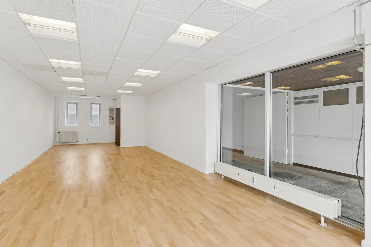 Store vinduer og åbne døre ud til gaden. Moderne design med hvidt loft og gulv. Lys lysekroner hængende fra loftet. Væggen er hvid og ren med enkelt radiator