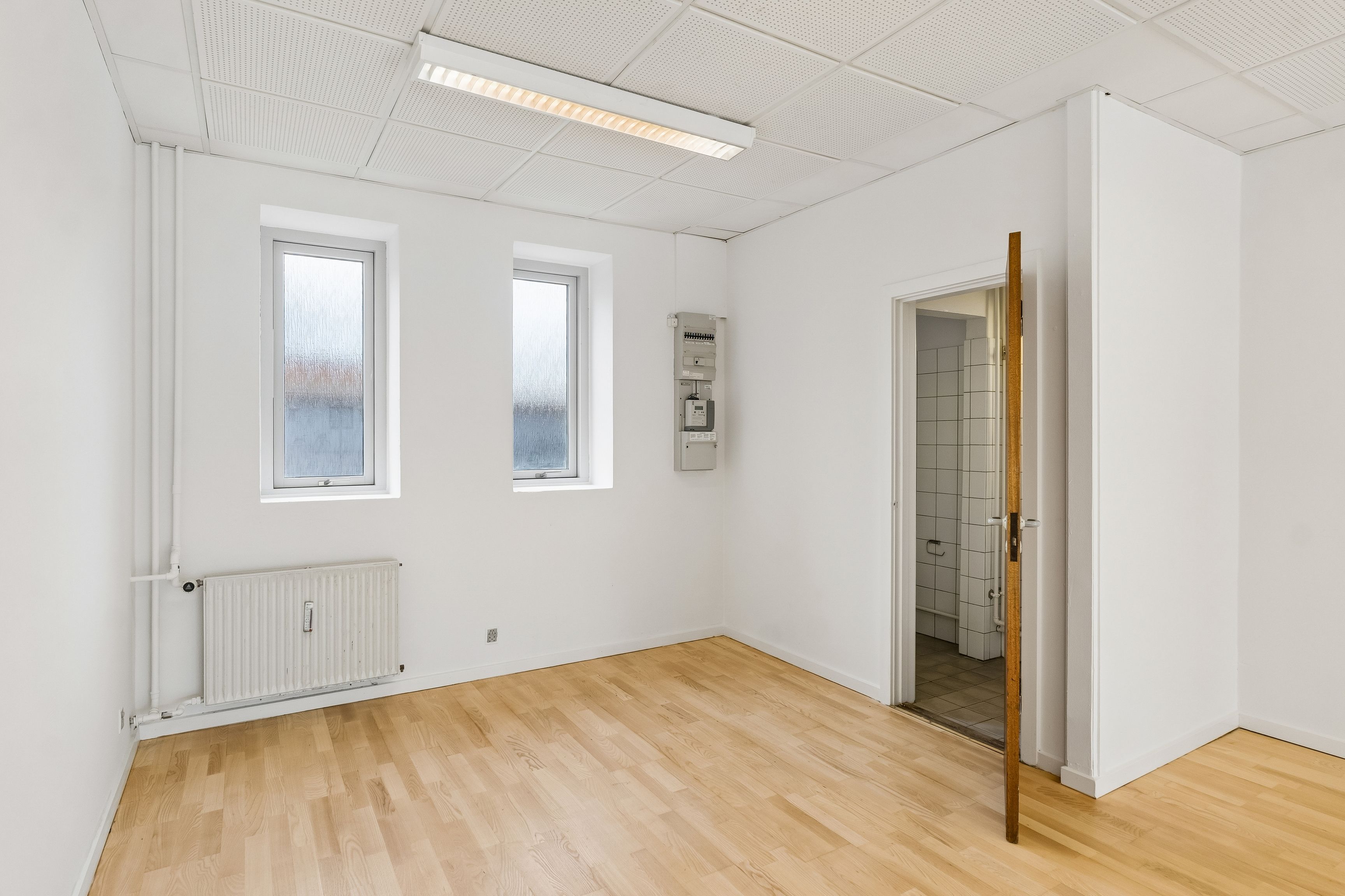 Store vinduer og højt loft. To vinduer, radiator og et lysegråt loftspanel. Trægulv og lysegrå vægge. Dør til et lille badeværelse og et åbent skab.