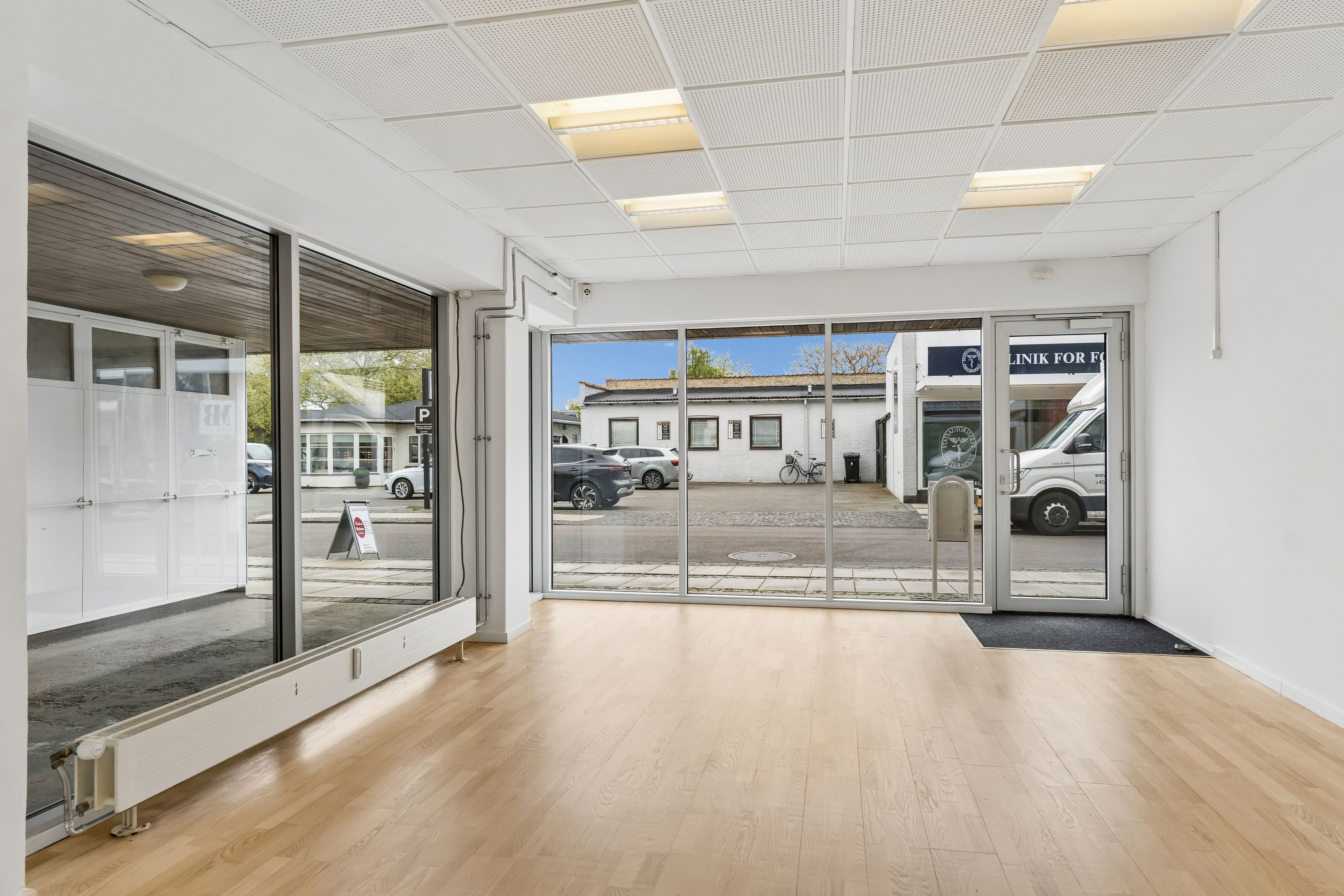 Foto af en butik med stort lyst vinduesfront og indgangsdøre i glas. På gaden udenfor er der parkerede biler, hvoraf en hvid er parkeret lige foran butikken