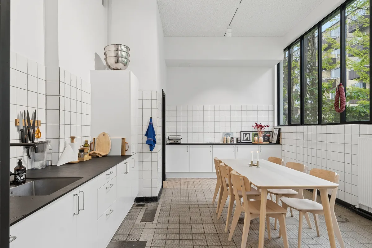 Minimalistisk køkken med lyse, hvide vægge og lysegrå bordplade samt lyse, lysegrå hjørneskabs. Væggene er klebet med hvide kakelister og lyse, hvide møbler