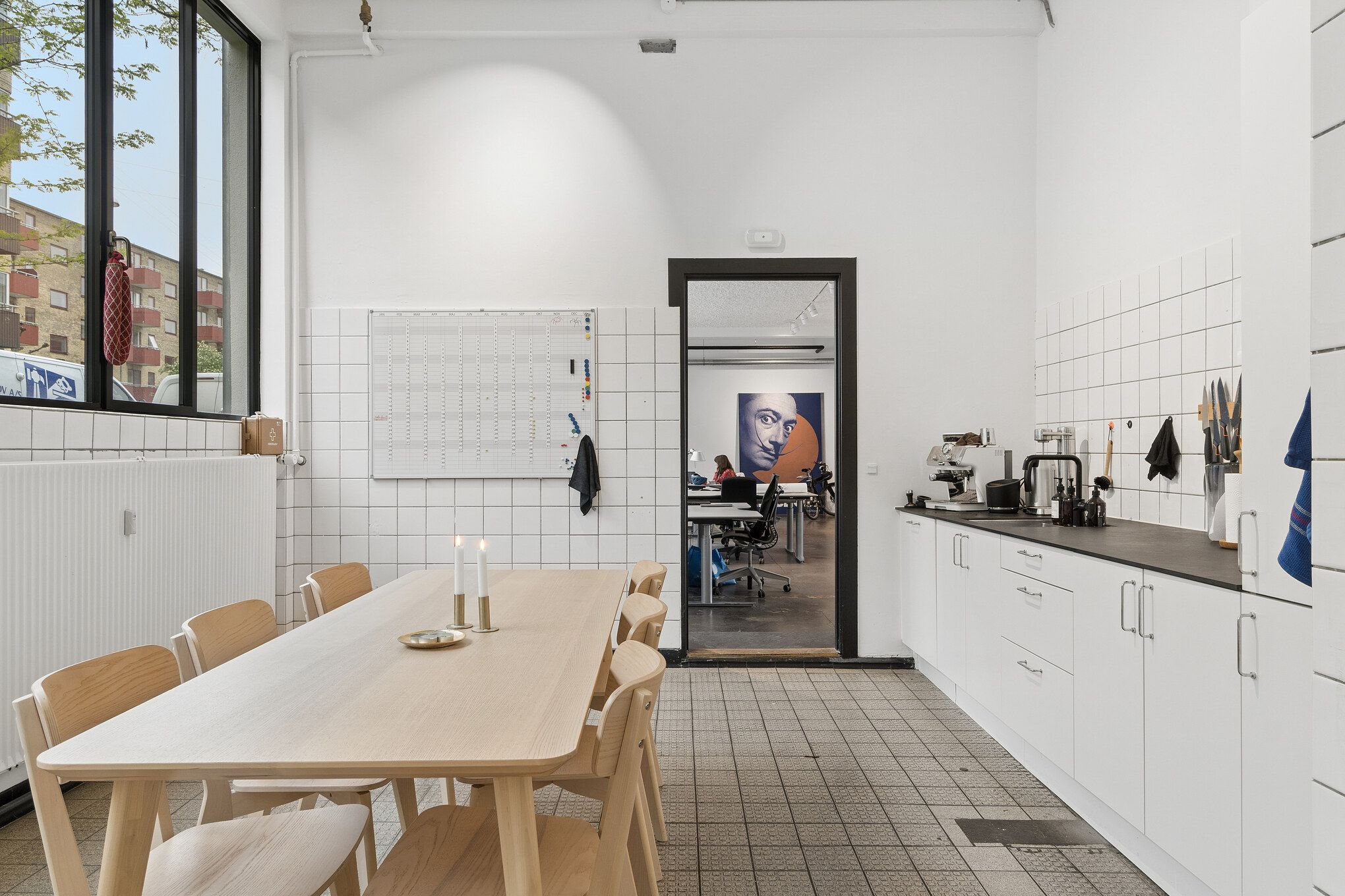 Kontorlokaler med minimalistisk indretning, hvor et bord med to lys, hvide køkkenskabe og en åben dør til et andet rum er synlige