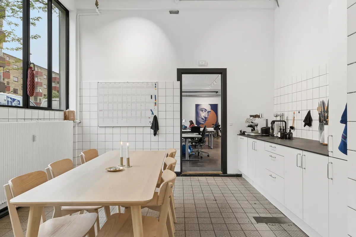Kontorlokaler med minimalistisk indretning, hvor et bord med to lys, hvide køkkenskabe og en åben dør til et andet rum er synlige