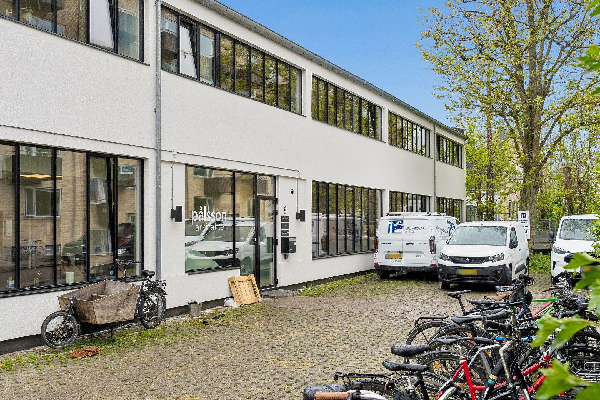 Hjørnebygning med glasvinduer og moderne design. Parkerede biler og cykler foran bygningen. Foran er der en stor butik med skiltet Paissøn.