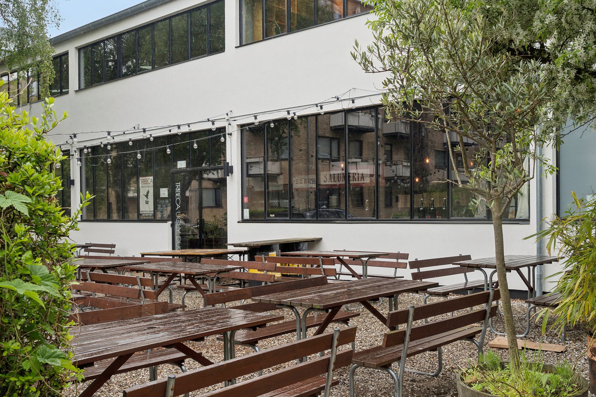 Værelser med terrasse og udsigt til grønne arealer og træer. Møbler er grupperet omkring spisesteder med borde og bænke. Kædelys er hængt over bordene.