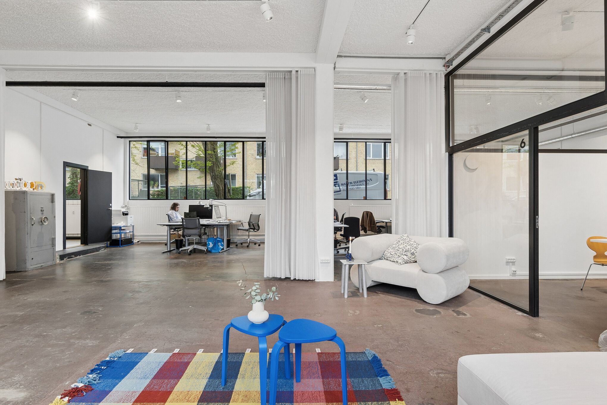 Kontorlokaler med åben planløsning og store vinduer. Stolene, skrivebord og sofaer med smuk tekstilbeklædning. Farverige gulvmåtter og moderne interiørdesign.