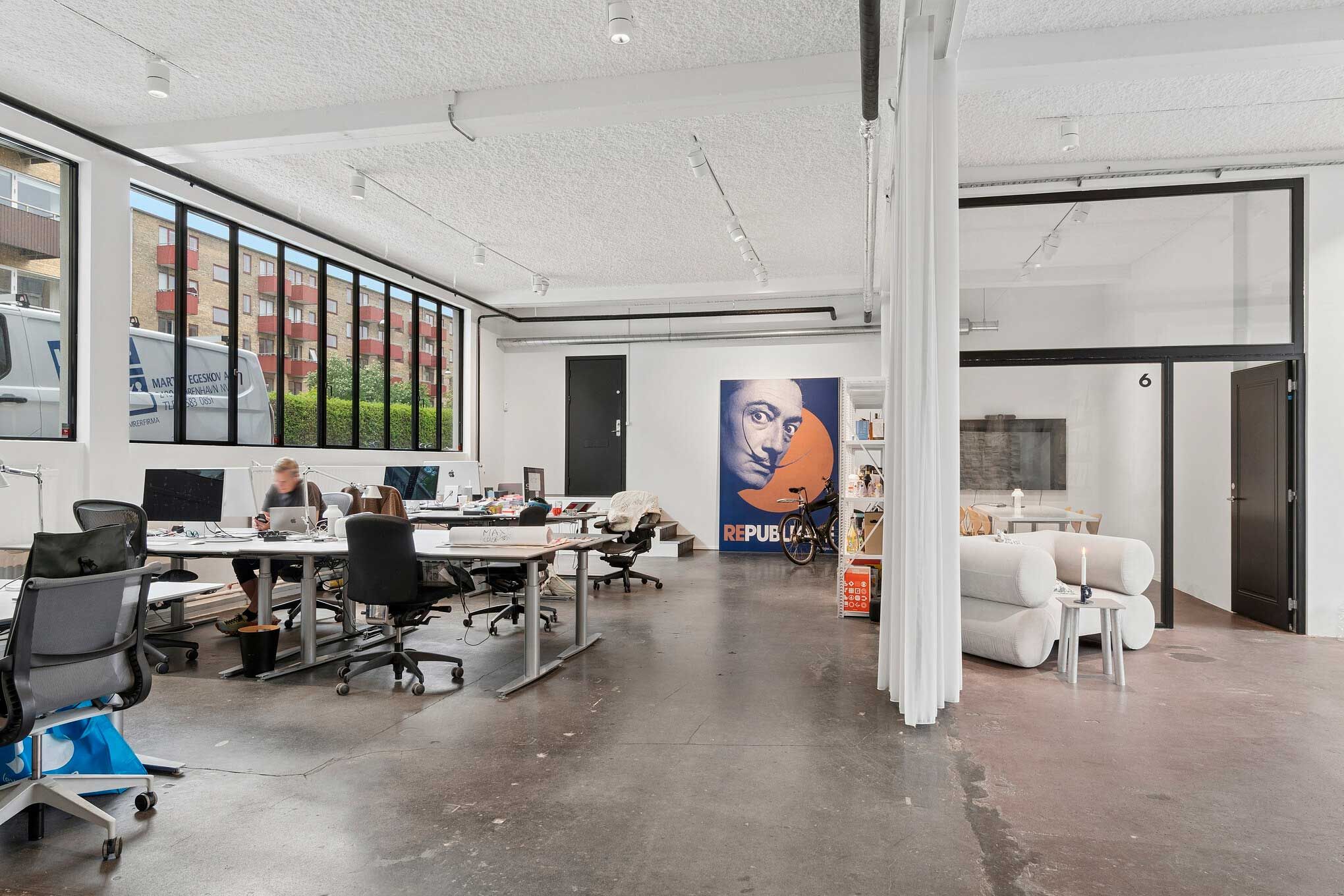Kontor med højloft, åbne arbejdsområder og højvinduer, hvor et team arbejder på bærbare og skriveborde med industriel charme