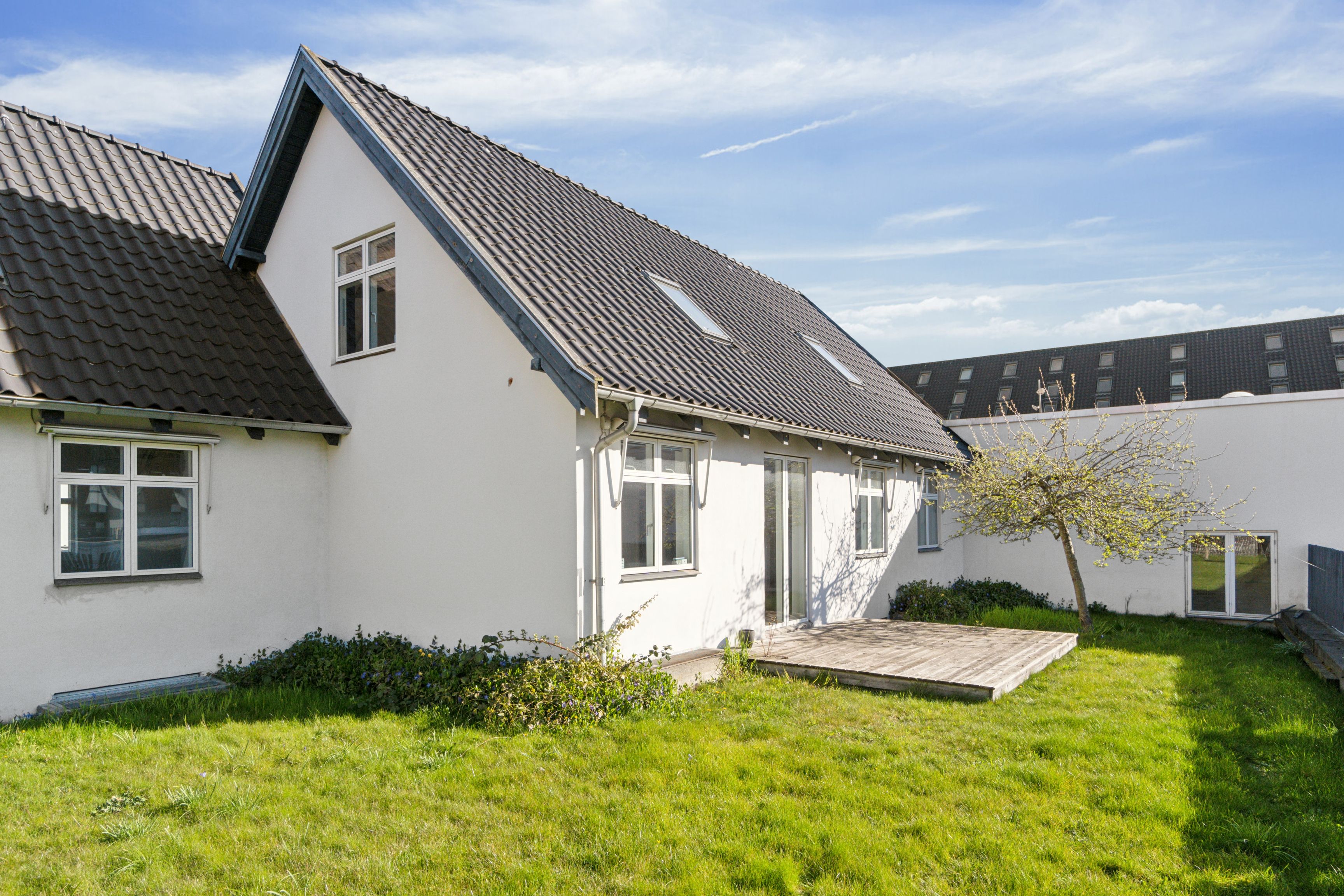 Hvidt malet kontorhuse med tagrør og terrasse, solrige ruder og grønt græs, slanke bygninger i baggrunden og lyse skyer på himlen