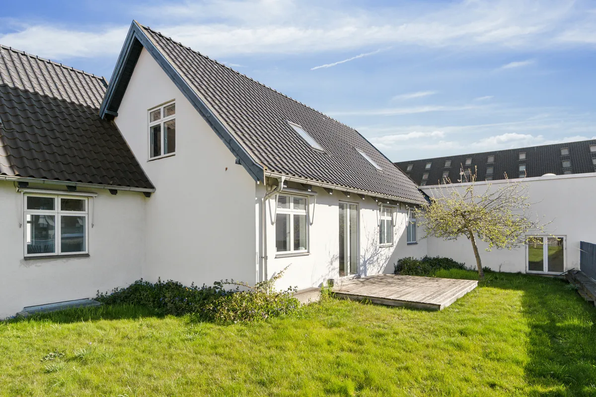 Hvidt malet kontorhuse med tagrør og terrasse, solrige ruder og grønt græs, slanke bygninger i baggrunden og lyse skyer på himlen