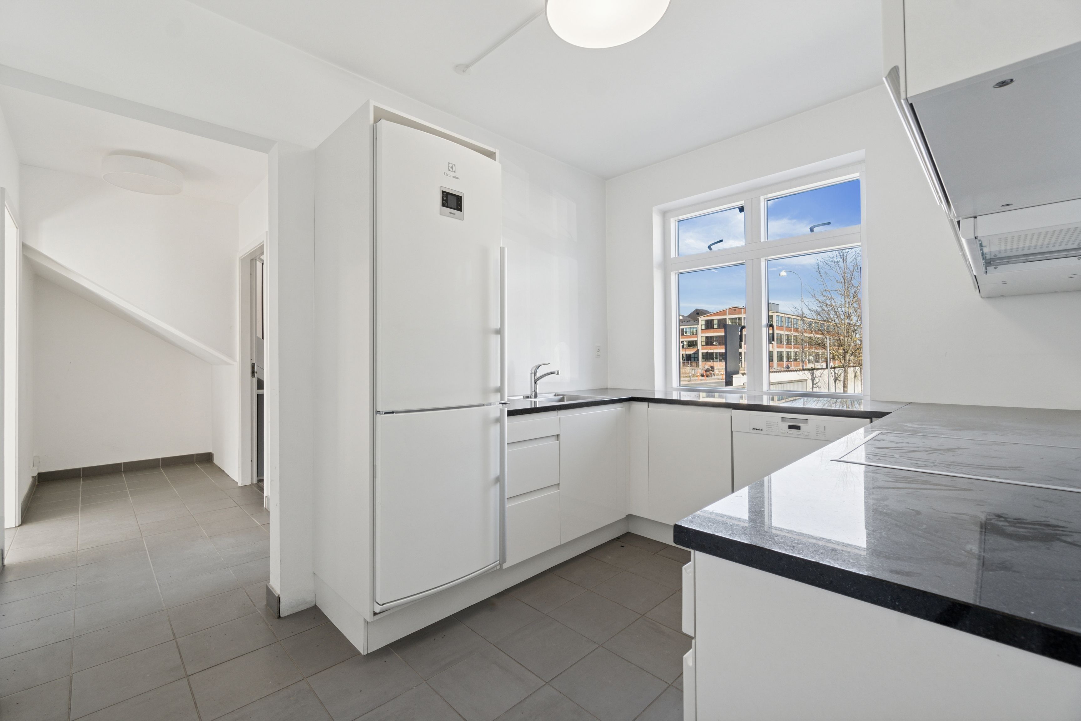 Hvidt moderne køkken med lys gulv og loft, lysekron, tekøkken med vaskemaskine, ovn og køleskab samt udgang til en have med terrasse.
