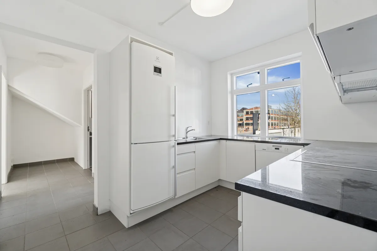 Hvidt moderne køkken med lys gulv og loft, lysekron, tekøkken med vaskemaskine, ovn og køleskab samt udgang til en have med terrasse.