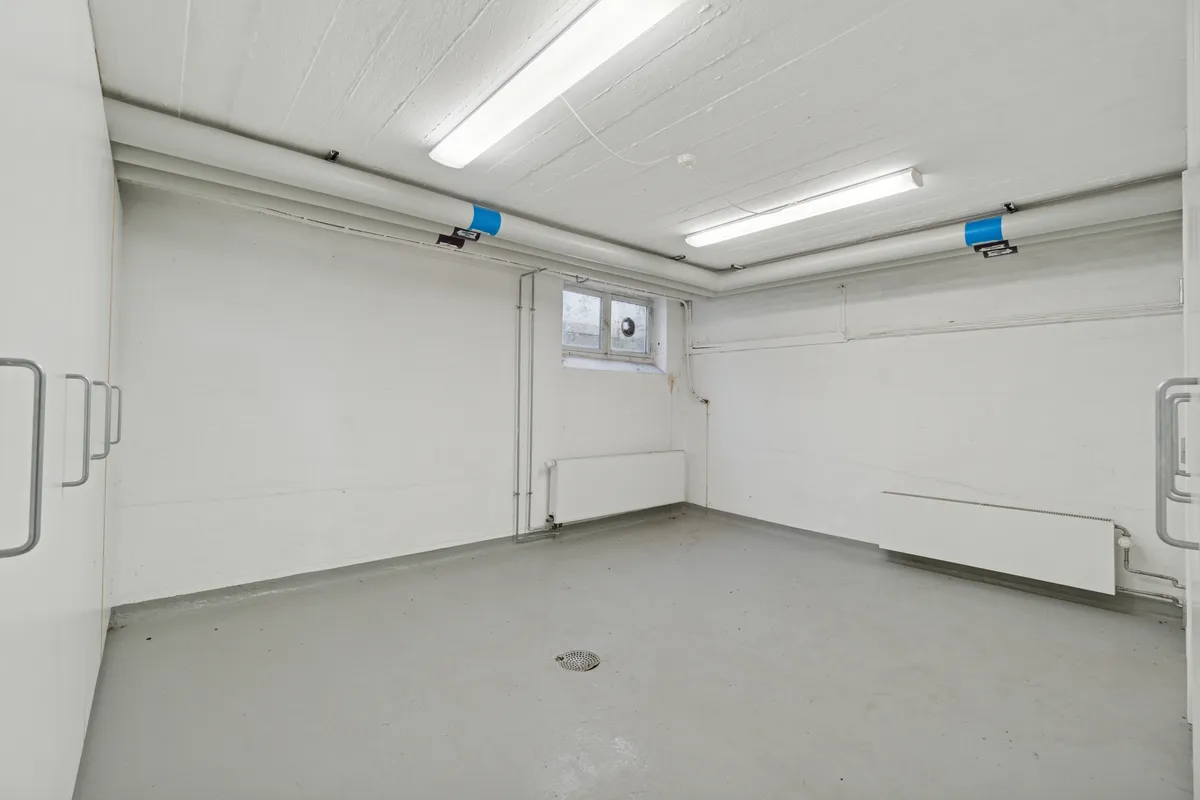 Et rum med hvide vægge og gulv, to radiatorer og to døre, samt to lyse rør på loftet.