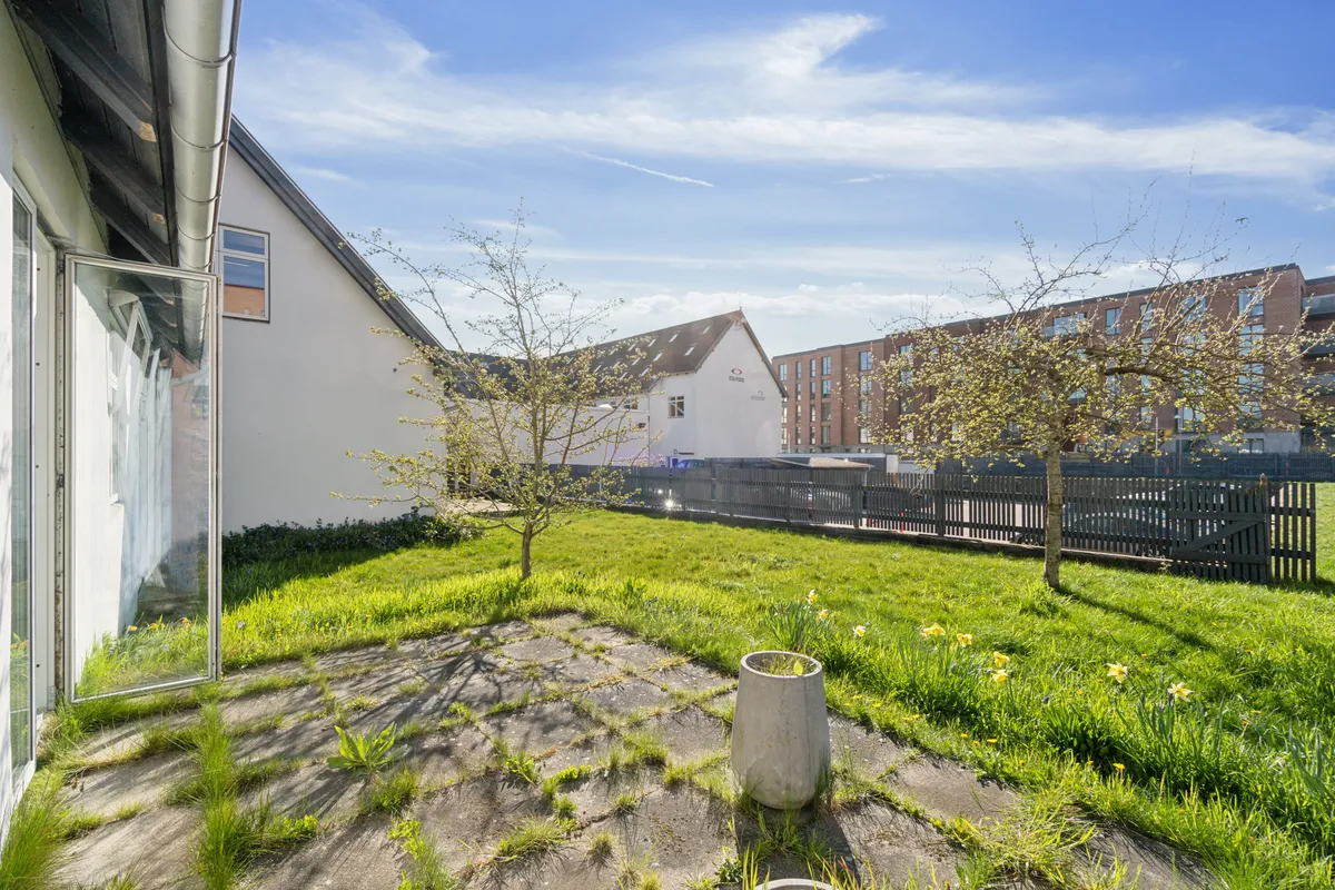 Mini-kontordomicil med terrasse ud mod en lille have, der er dækket af græs og små blomster. Foran er et skur med glasdør og et cementstøbt skulptur.